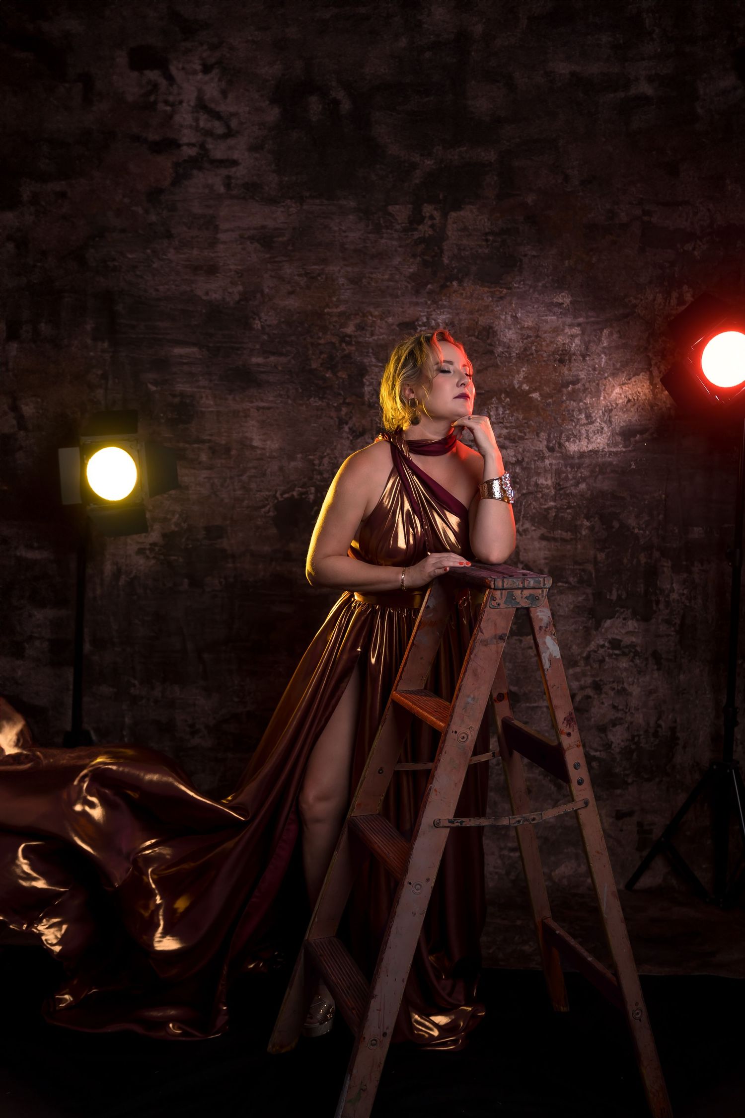 A dramatic studio photo with a woman in a flowing brown dress posed against a dark wall with yellow and red spotlights.