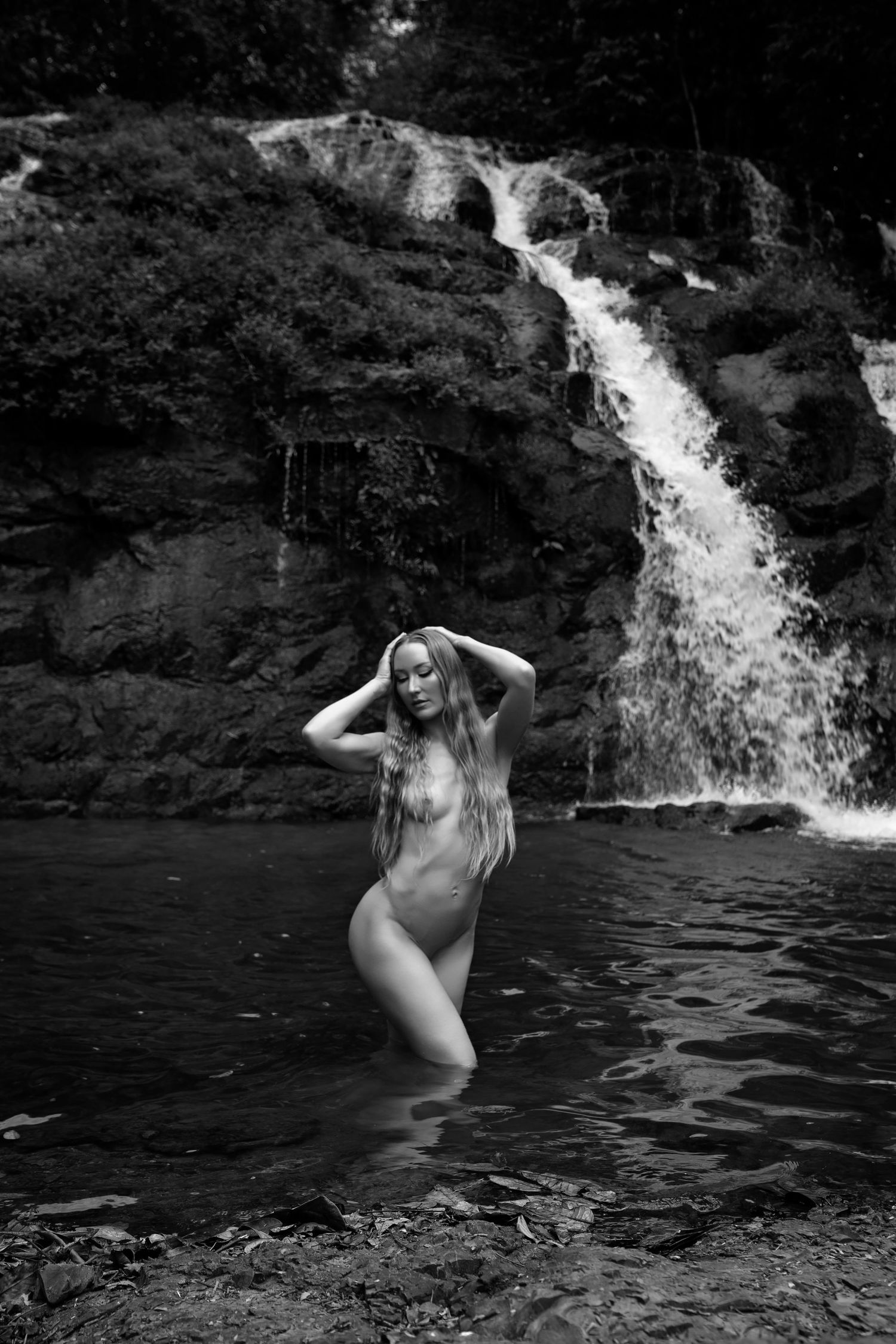 Black and white photo of a woman standing in a natural pool before a cascading waterfall surrounded by rocks.