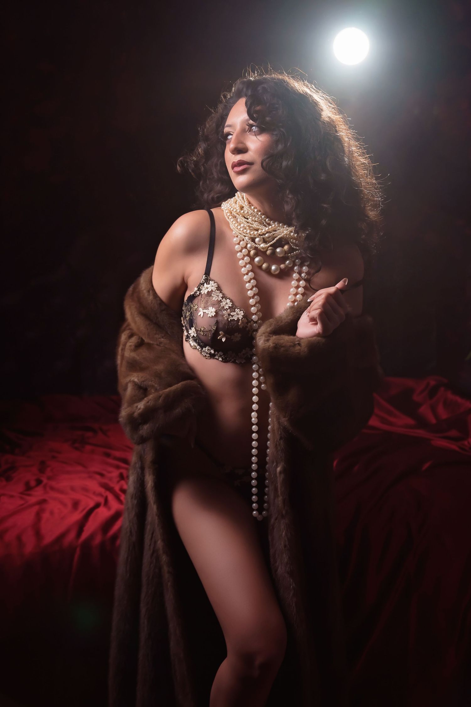 Dramatic backlit boudoir photo with model in pearls and fur coat on red backdrop.