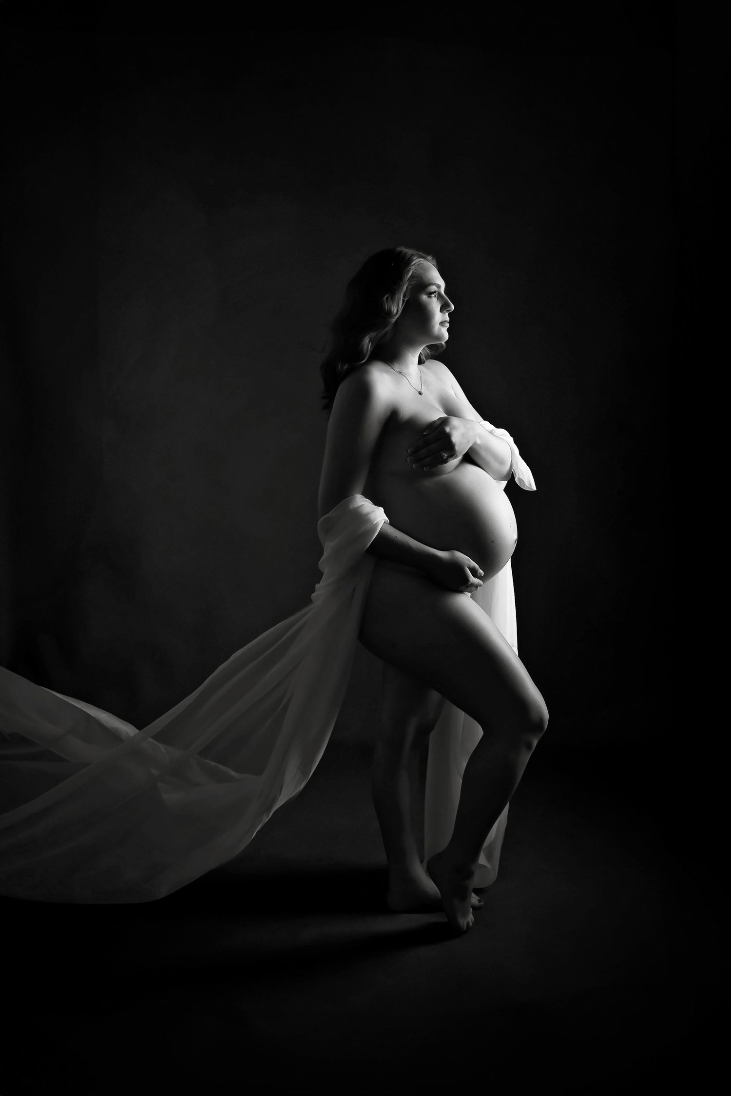 Low key maternity photography with dramatic lighting and flowing fabric elements.