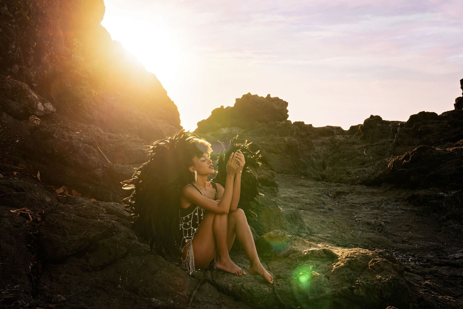 Woman sitting on rocky shore at golden sunset, bathed in warm sunlight with ocean waves in background.