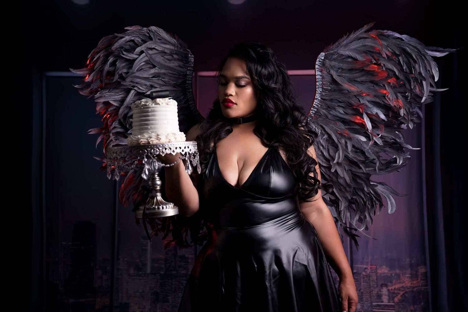 Woman in black latex dress with dark angel wings holds white cake in dramatic dark studio setting.