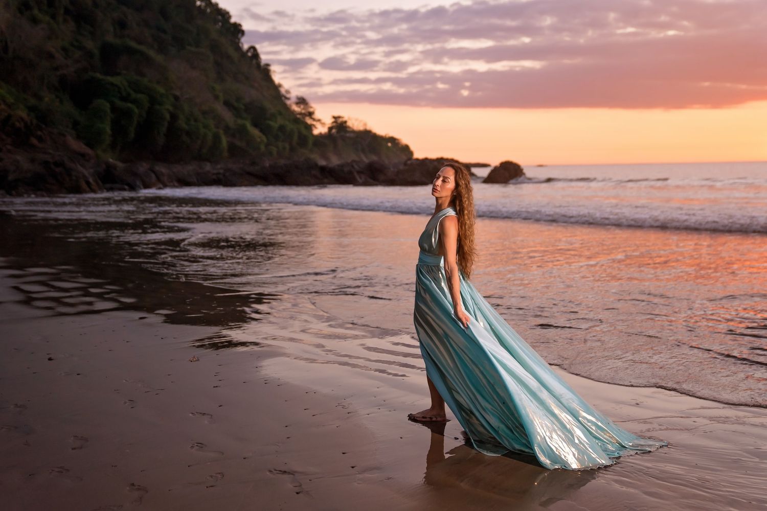 Woman in flowing teal gown standing on beach at sunset with waves and dramatic pink sky backdrop.