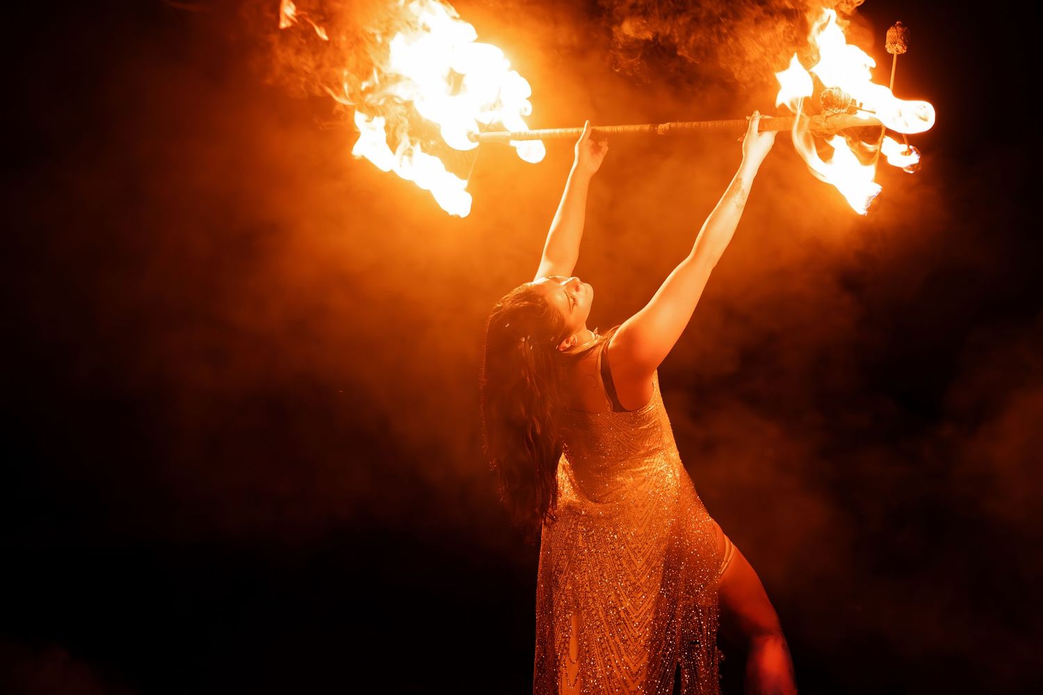 A fire performer holds flaming torches aloft against a dark smoky night sky, illuminated by dramatic orange flames.