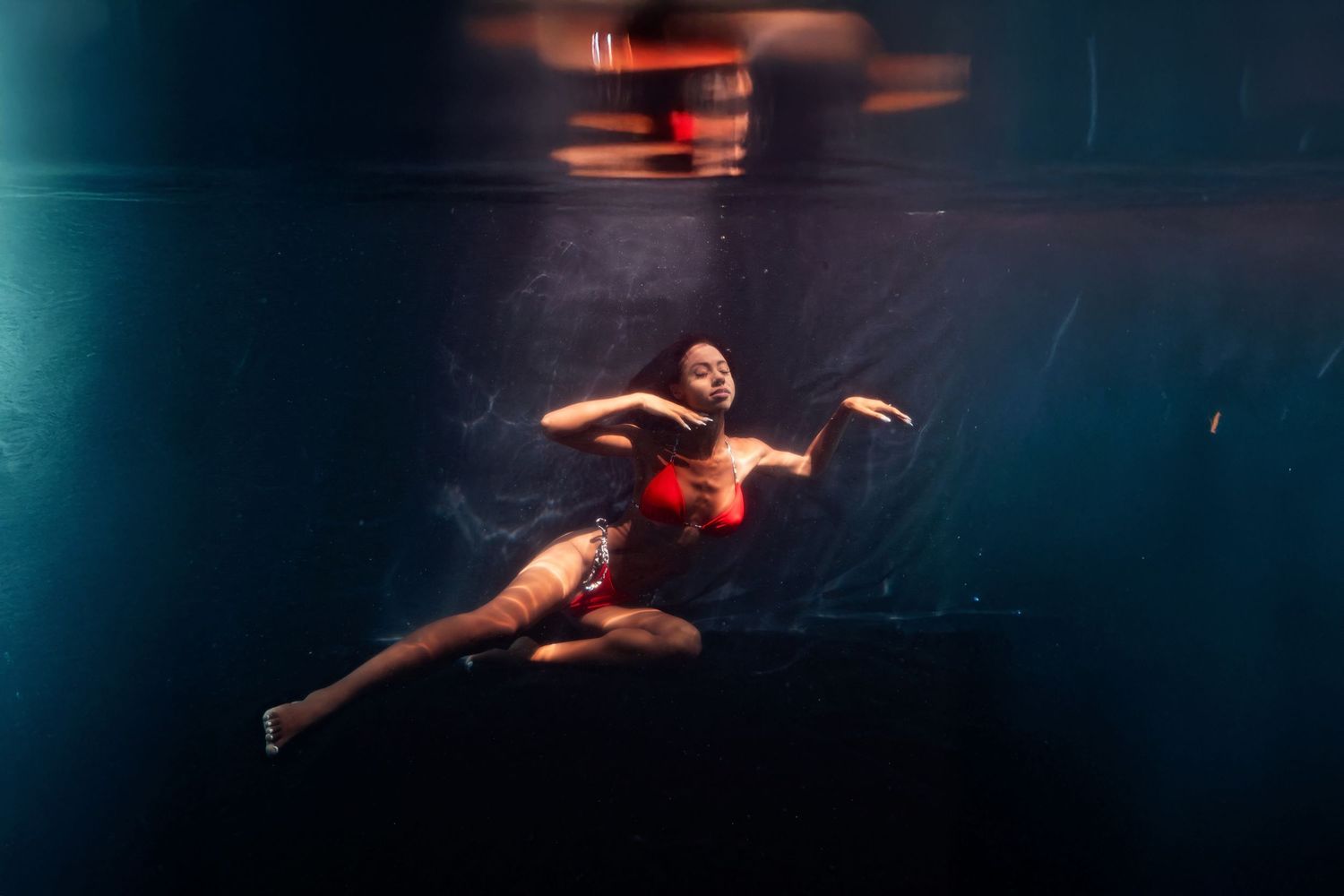 Woman in red bikini gracefully floating underwater in a dark pool with dramatic blue lighting.