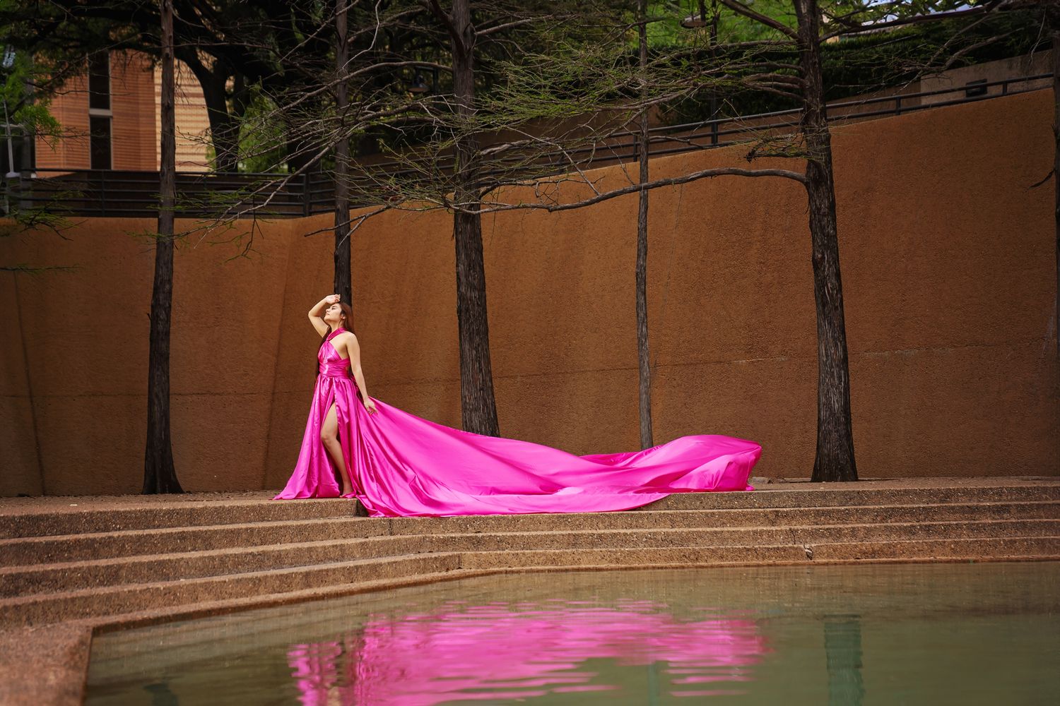 A woman in a flowing hot pink gown stands near a reflecting pool with tall trees and a terracotta wall in the background.