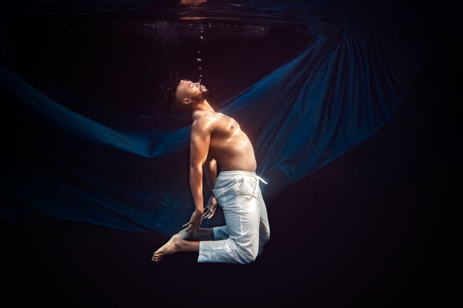 Figure suspended underwater against dark background with flowing blue fabric creates ethereal underwater dance scene.
