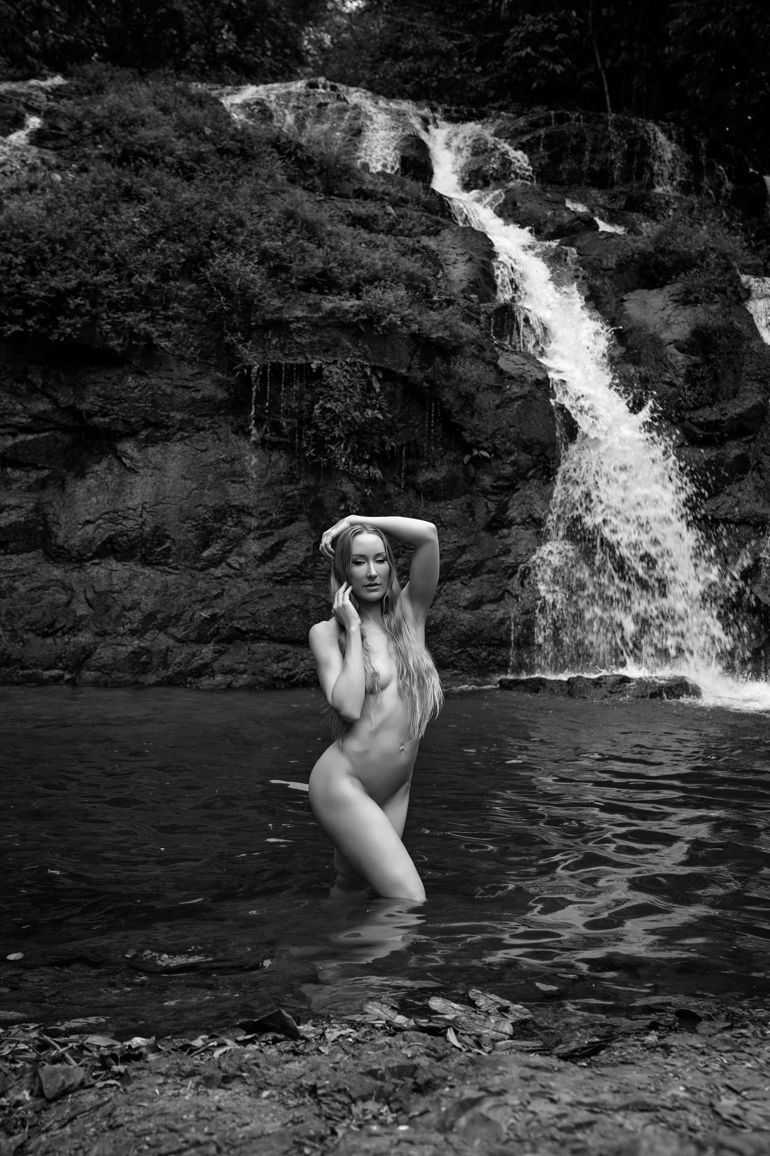 Black and white photo of a woman standing in a natural pool before a cascading waterfall surrounded by lush rocks.