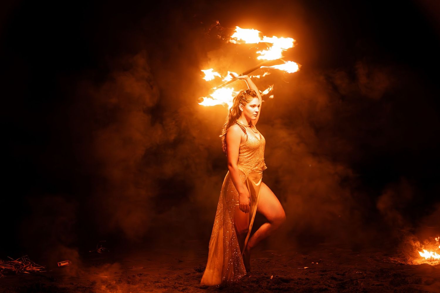 Woman in flowing dress performing fire dancing at night with flames illuminating the smoky scene.
