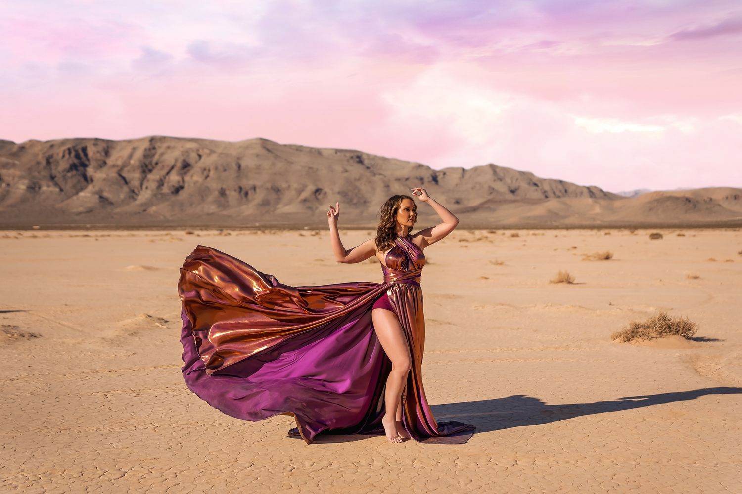 A flowing burgundy dress billows dramatically in the wind against a desert landscape with mountains during sunset.