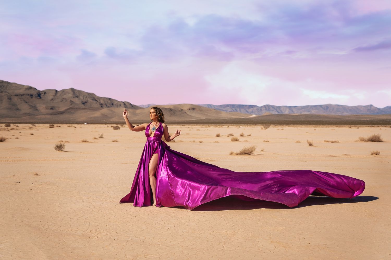 A flowing purple gown on a woman billows gracefully across a vast desert landscape at sunset.