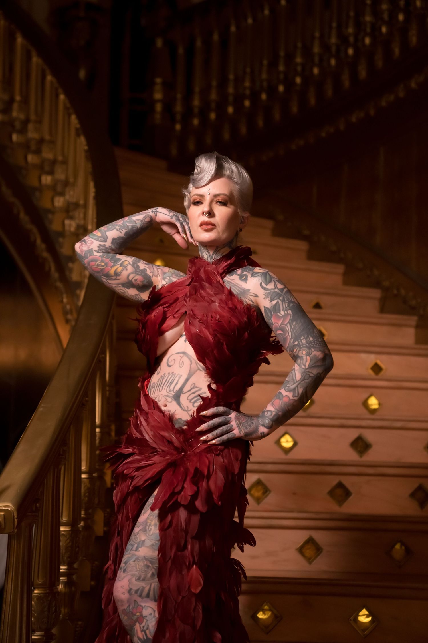 Tattooed woman in red feather gown poses elegantly on ornate wooden staircase.