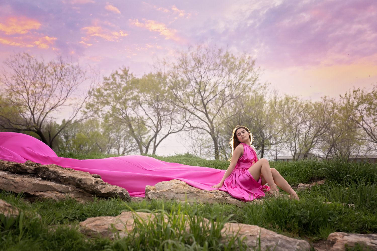 A flowing pink dress creates an ethereal effect against a moody sky with trees at sunset in a springtime landscape.