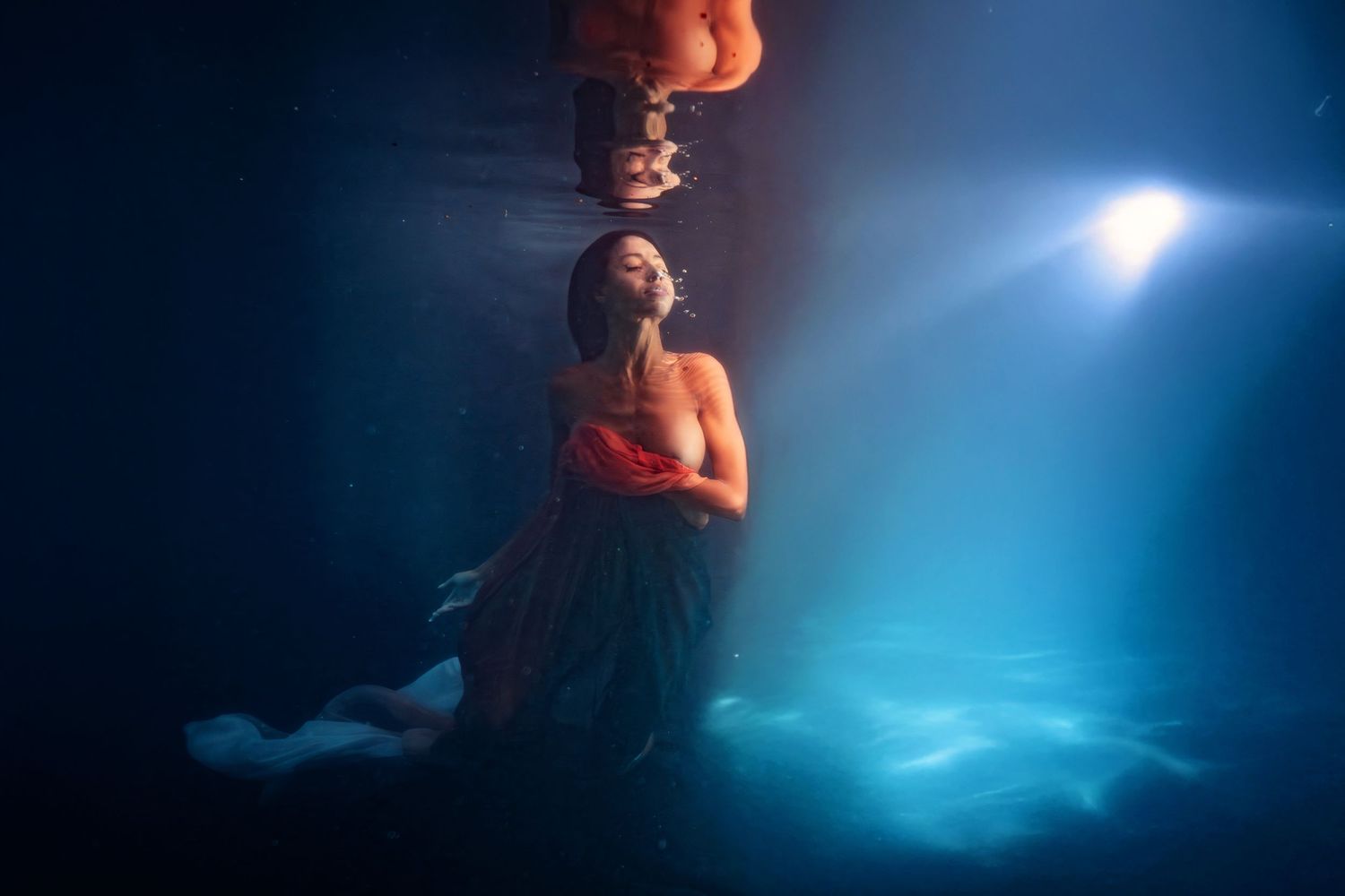 Woman in red top and dark skirt floating underwater with blue light rays.