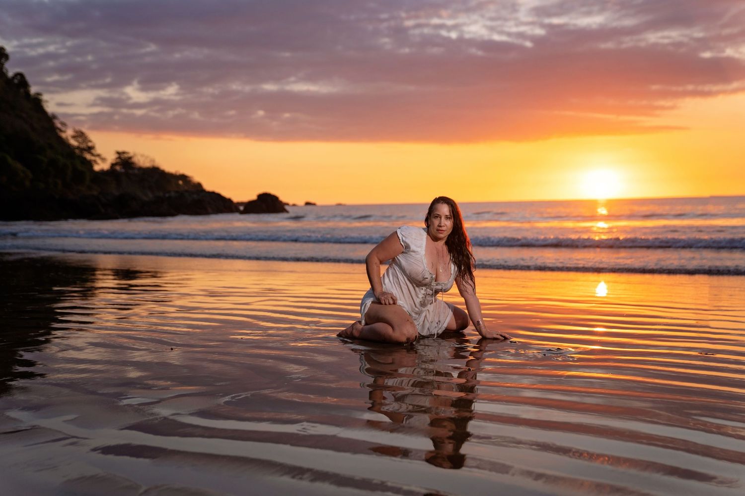 Woman in white dress sitting on a wet beach at golden sunset with dramatic sky and ocean waves.