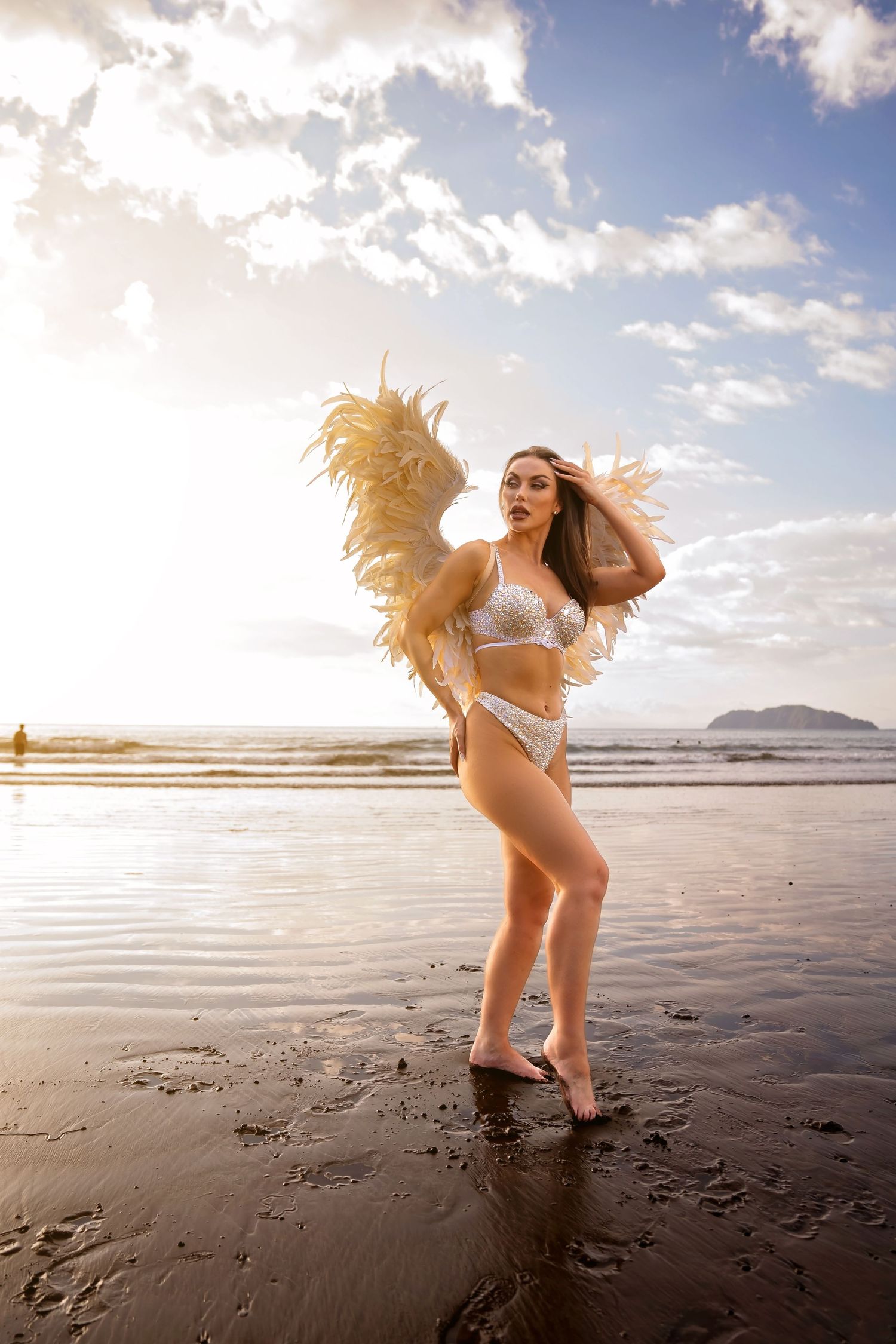 Woman in white sequin bikini with feather wings poses on a wet beach at golden sunset hour.