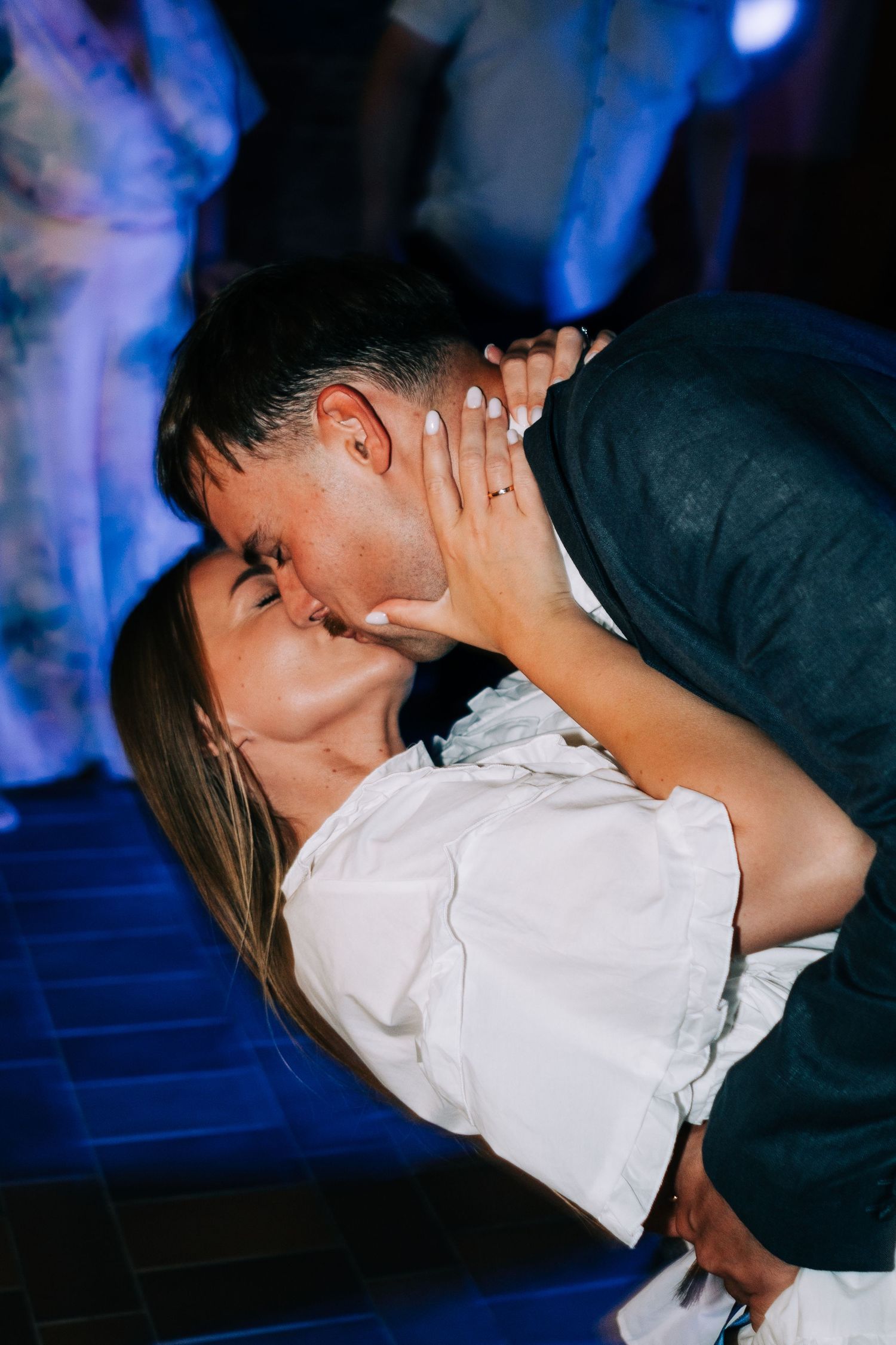 A romantic couple in formal attire sharing a passionate kiss against a dark blue-lit background.