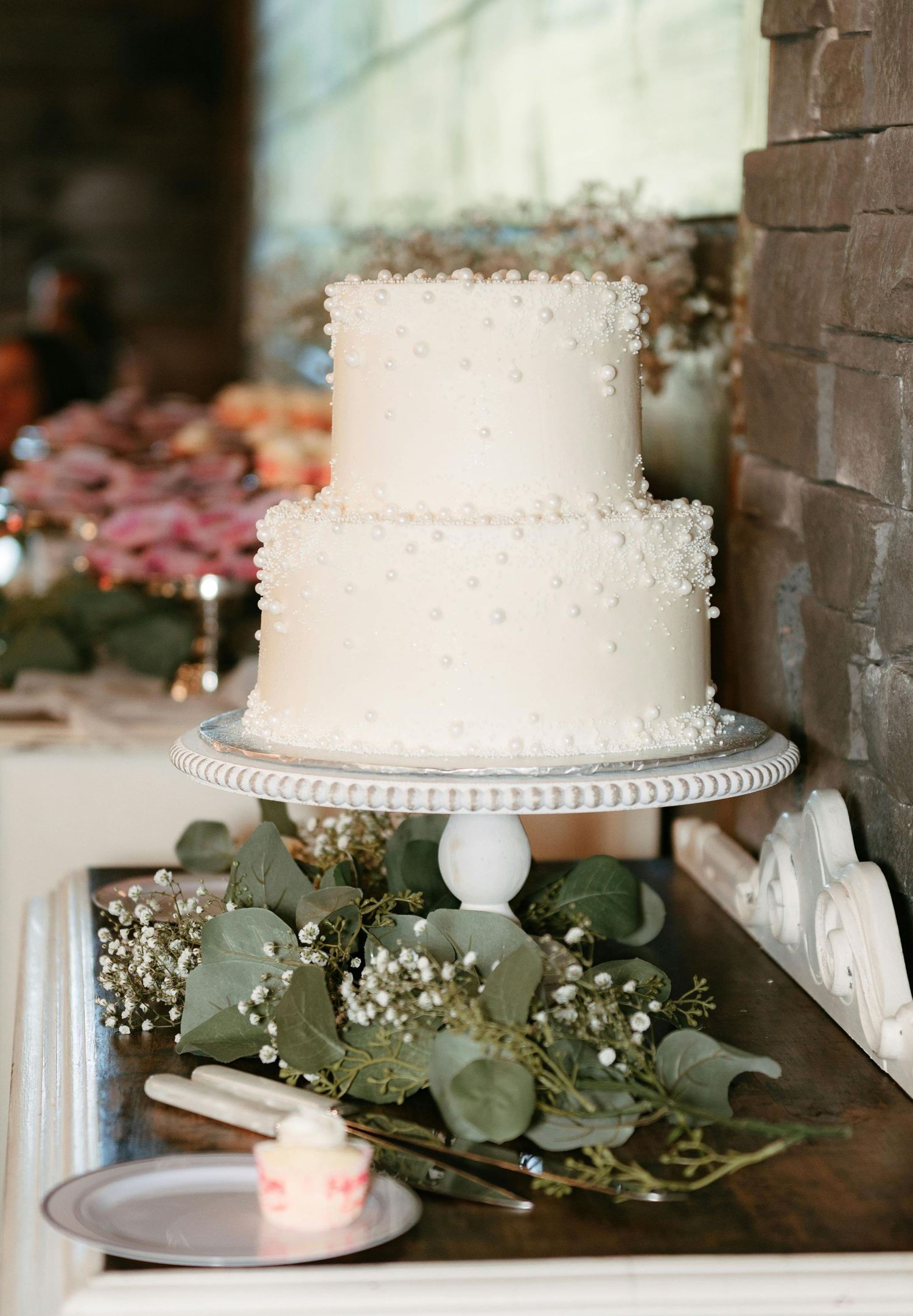 White tiered wedding cake with delicate pearl details displayed on elegant stands with eucalyptus greenery decorations.