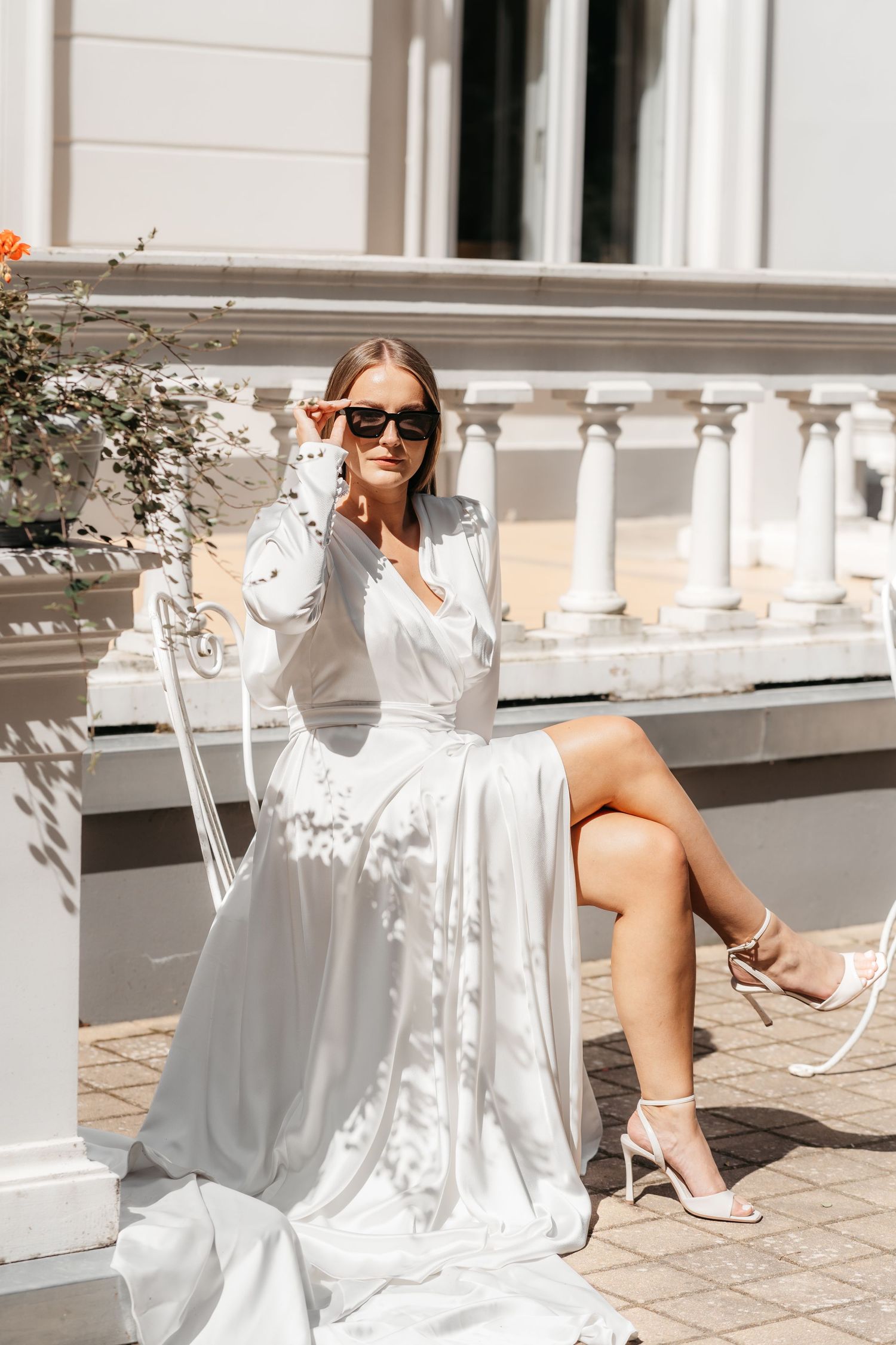 Woman in flowing white dress lounges elegantly on outdoor patio with dappled sunlight and architectural details.