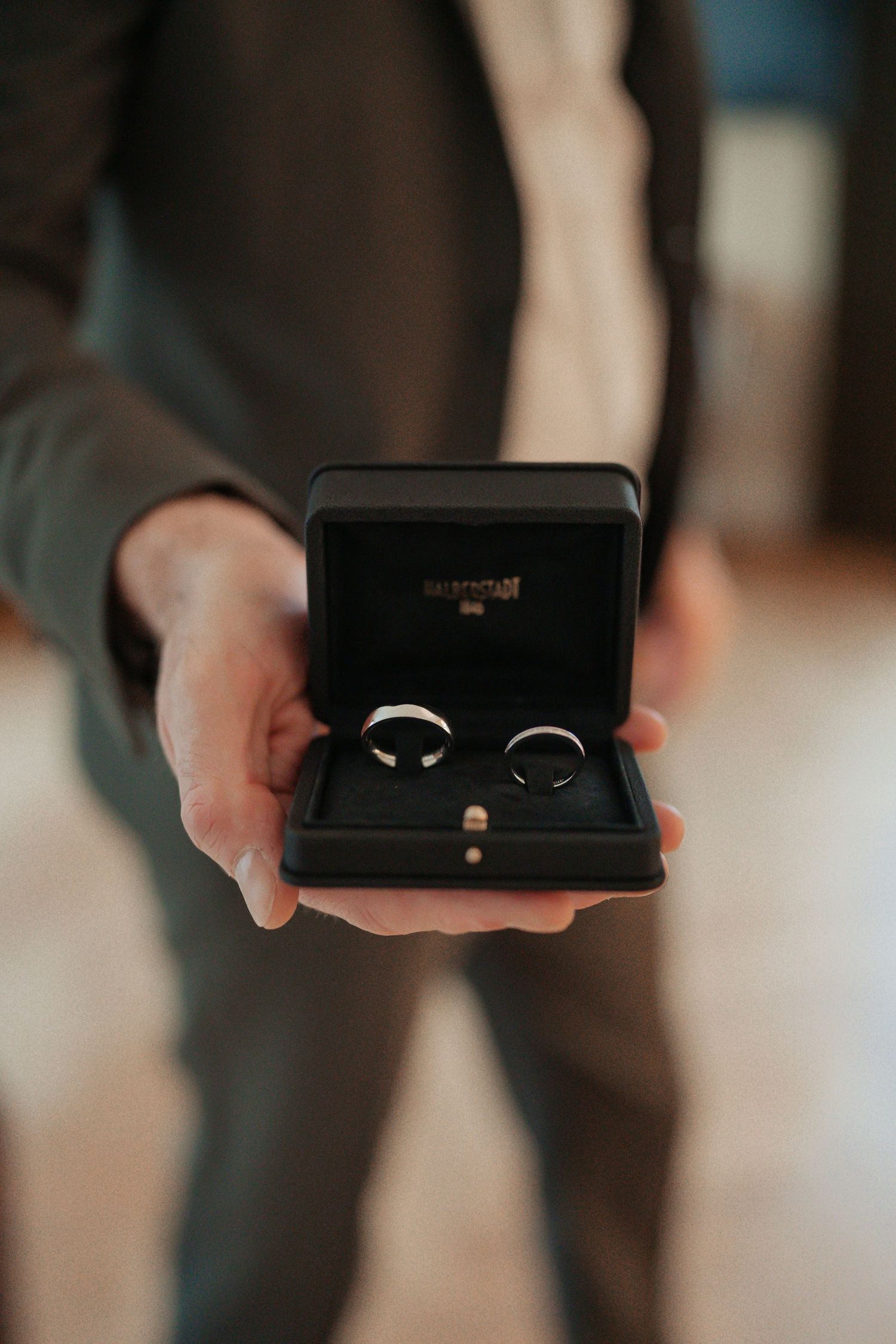 Hands presenting an opened black ring box with wedding band.