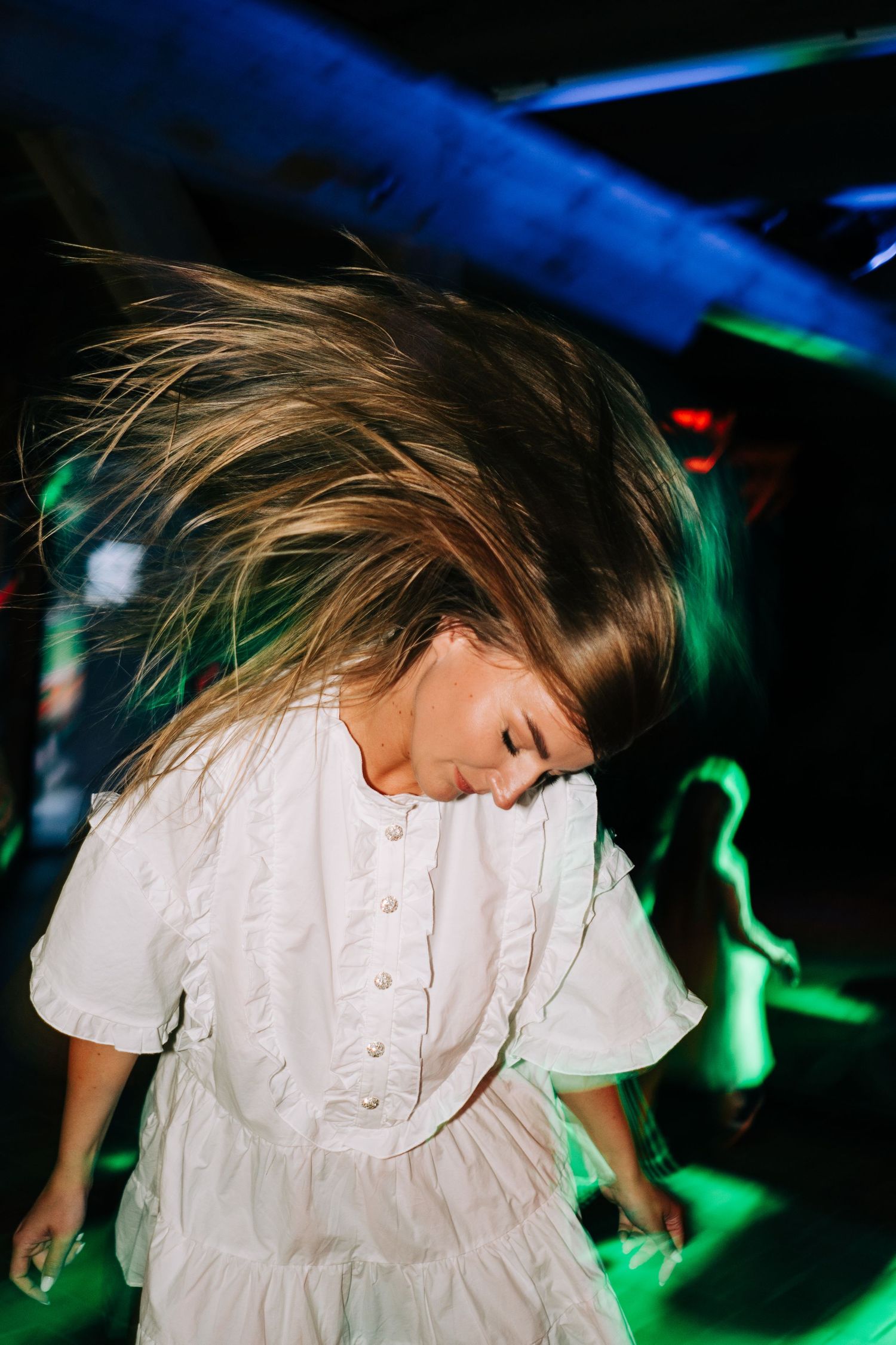 Person in white dress dancing energetically under blue and green party lights.