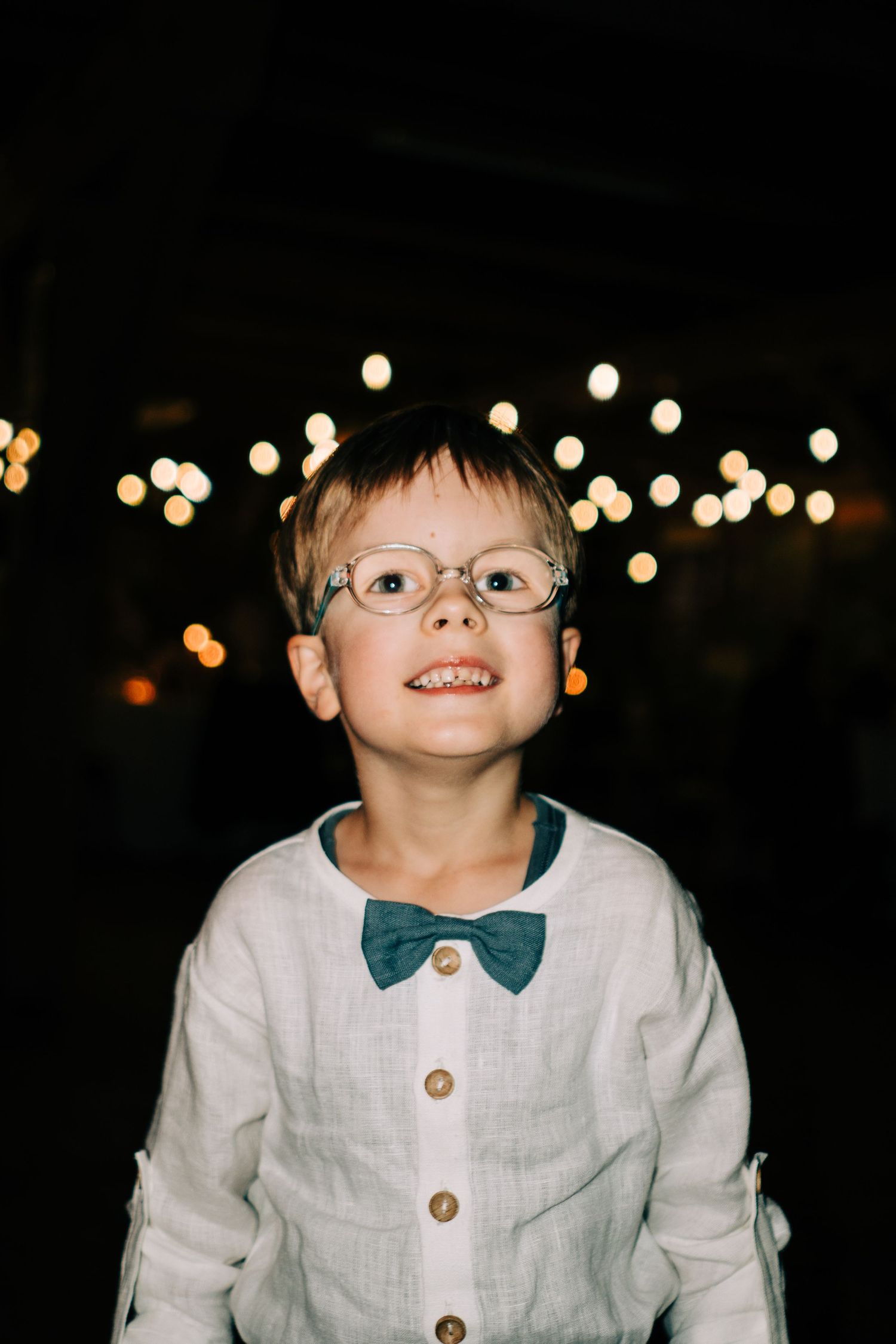 Child wearing white linen suit with bow tie smiles in front of bokeh lights at night.