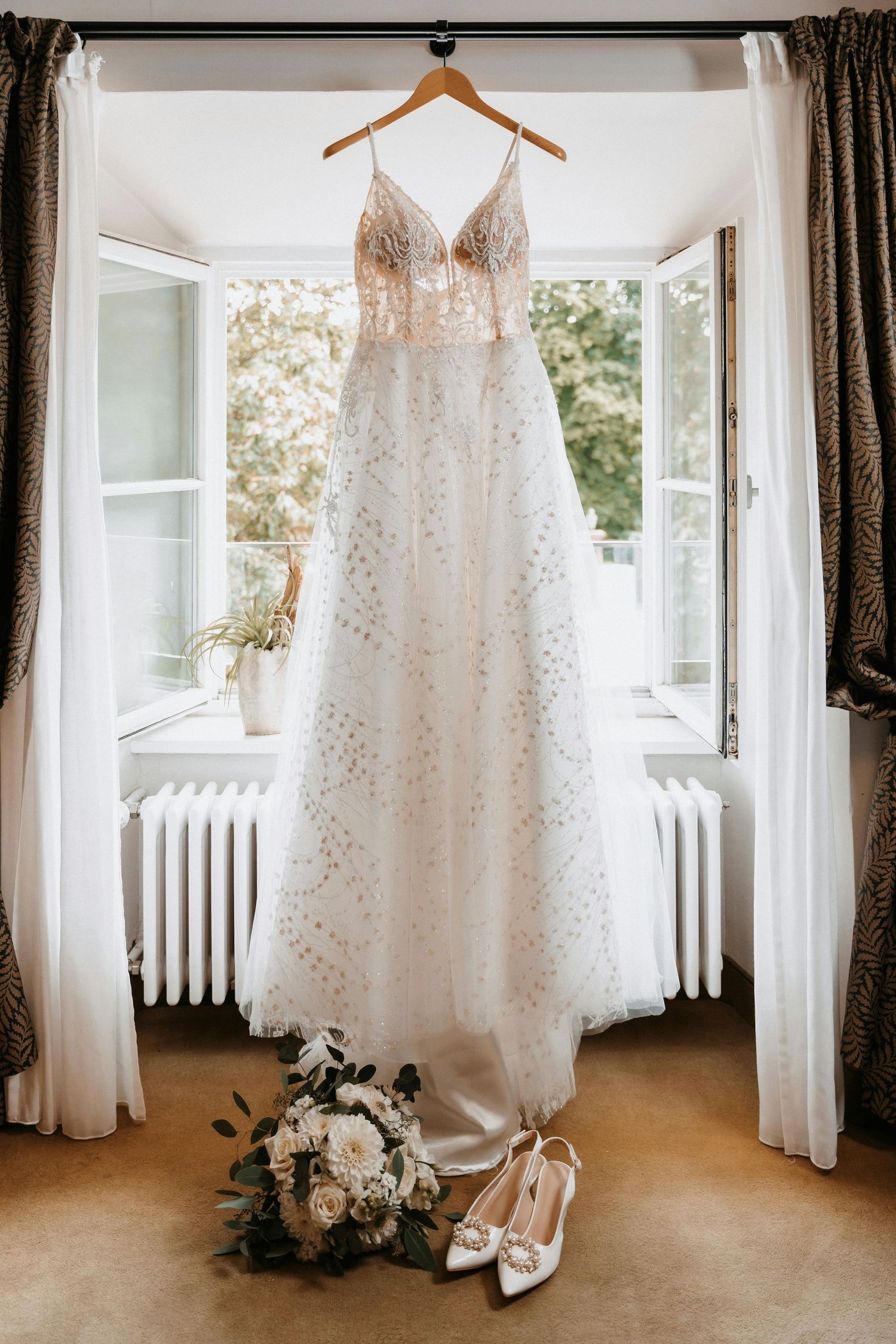 Elegant lace wedding dress hanging in window frame with bouquet and shoes below.