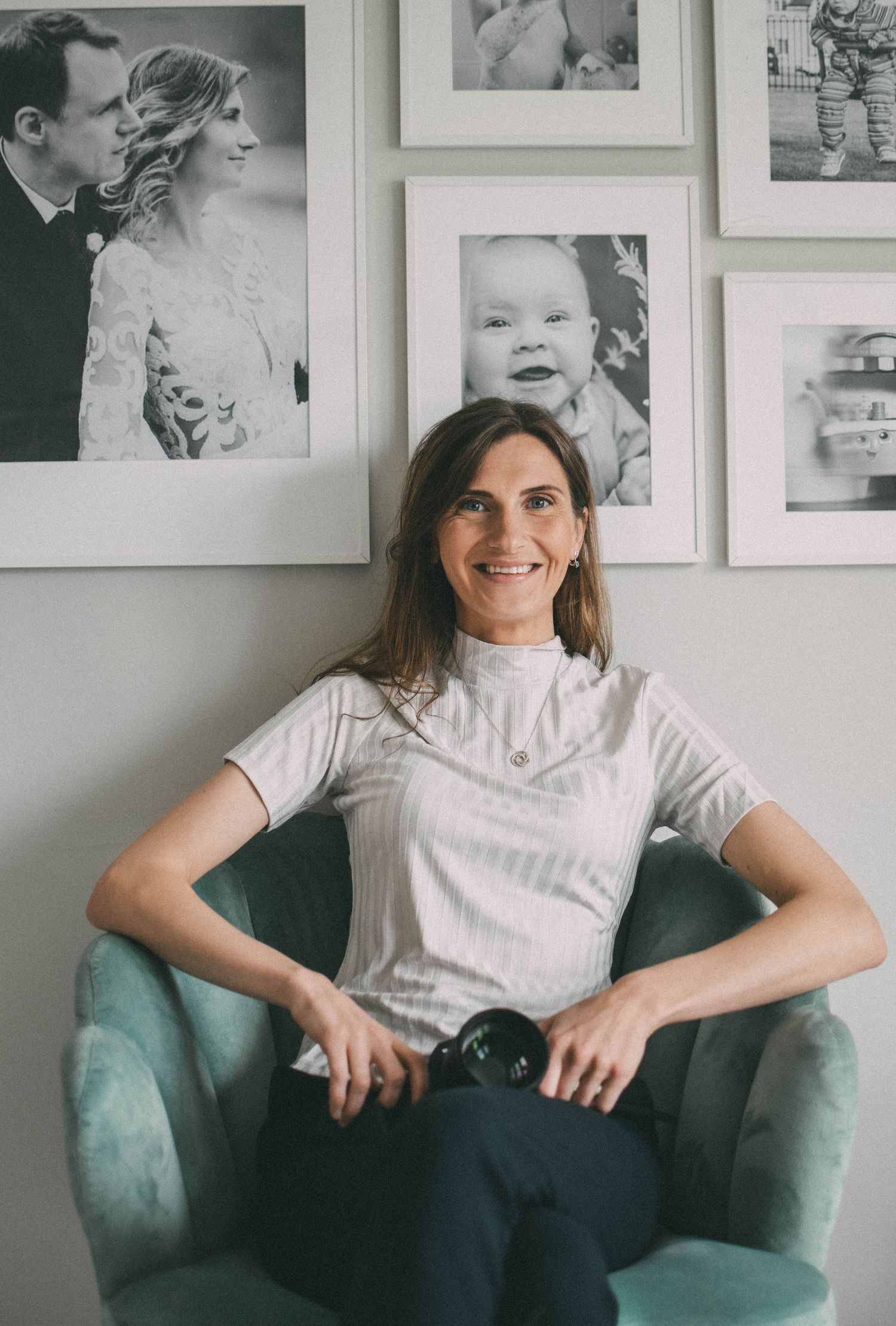 Person relaxed in a teal armchair wearing casual white shirt and dark pants against a wall of framed photos.
