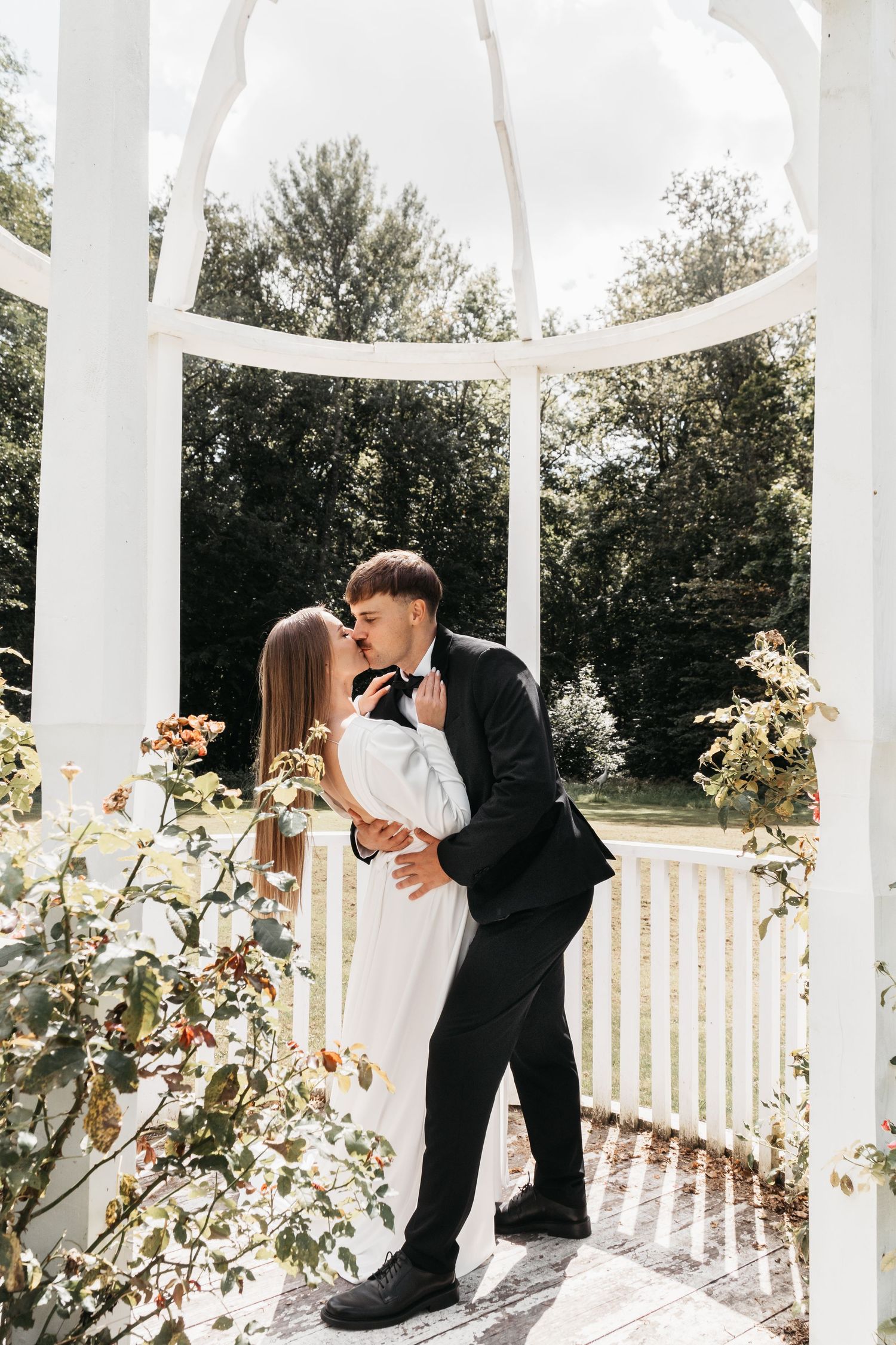 A romantic wedding kiss between newlyweds under an elegant white gazebo surrounded by blooming flowers.