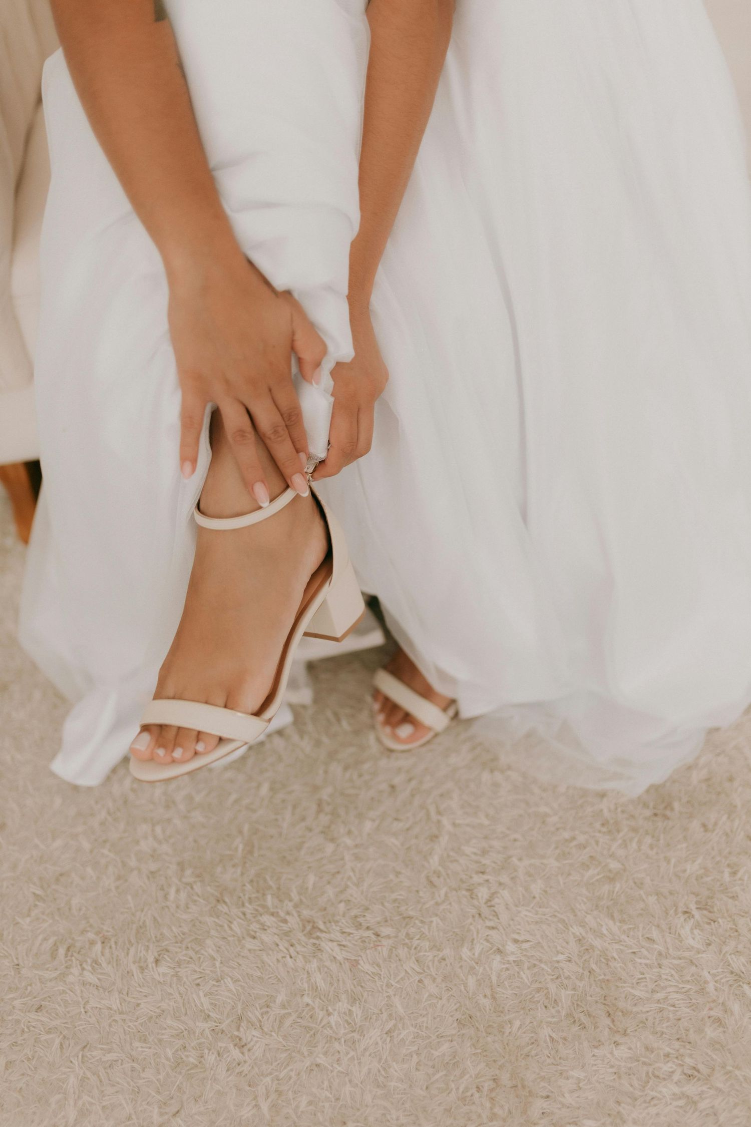 Close-up of hands adjusting white wedding shoes with delicate ankle straps against a white dress.