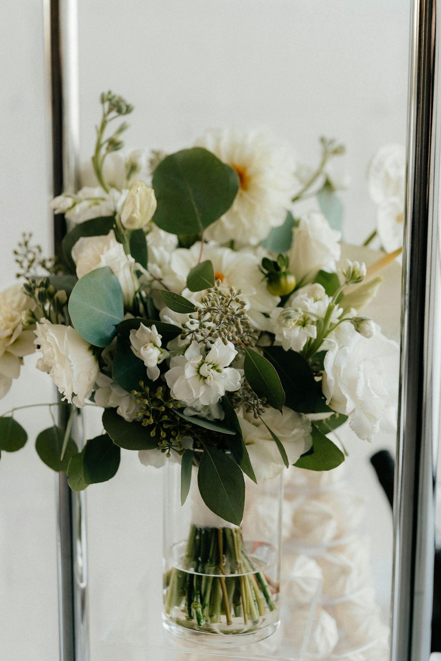 A delicate white floral arrangement with greenery in a clear glass vase features roses, dahlias and eucalyptus leaves.