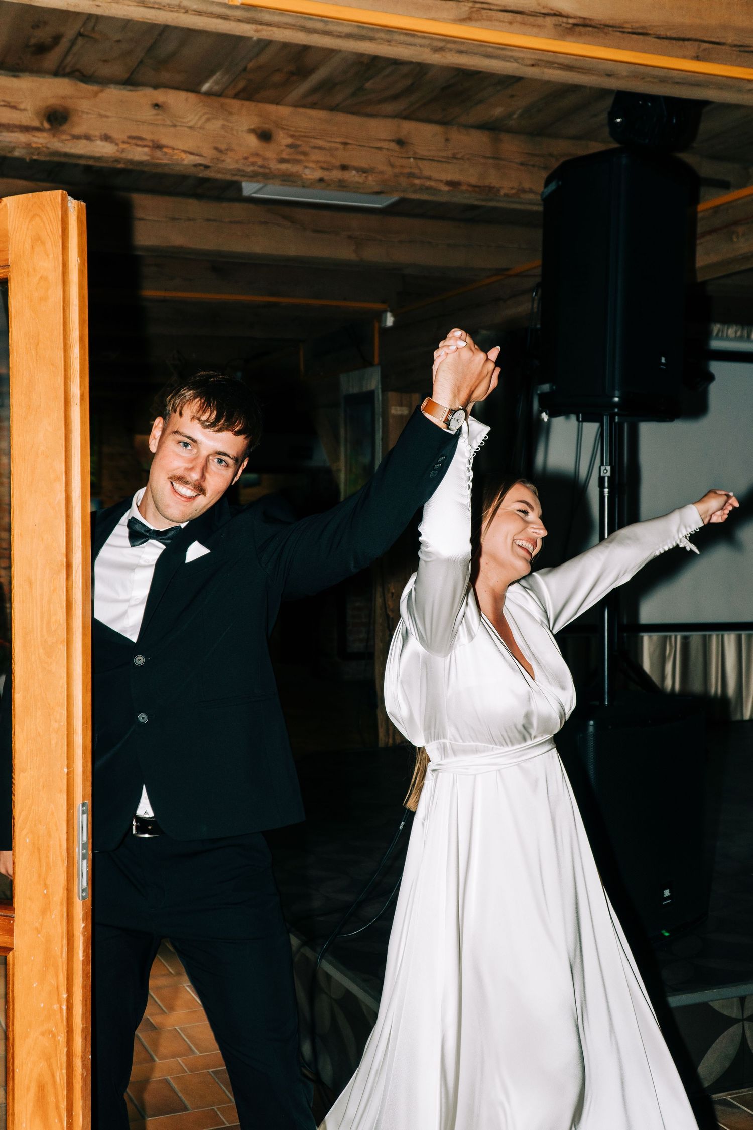 A couple in formal wedding attire dance joyfully together in a rustic wooden venue.