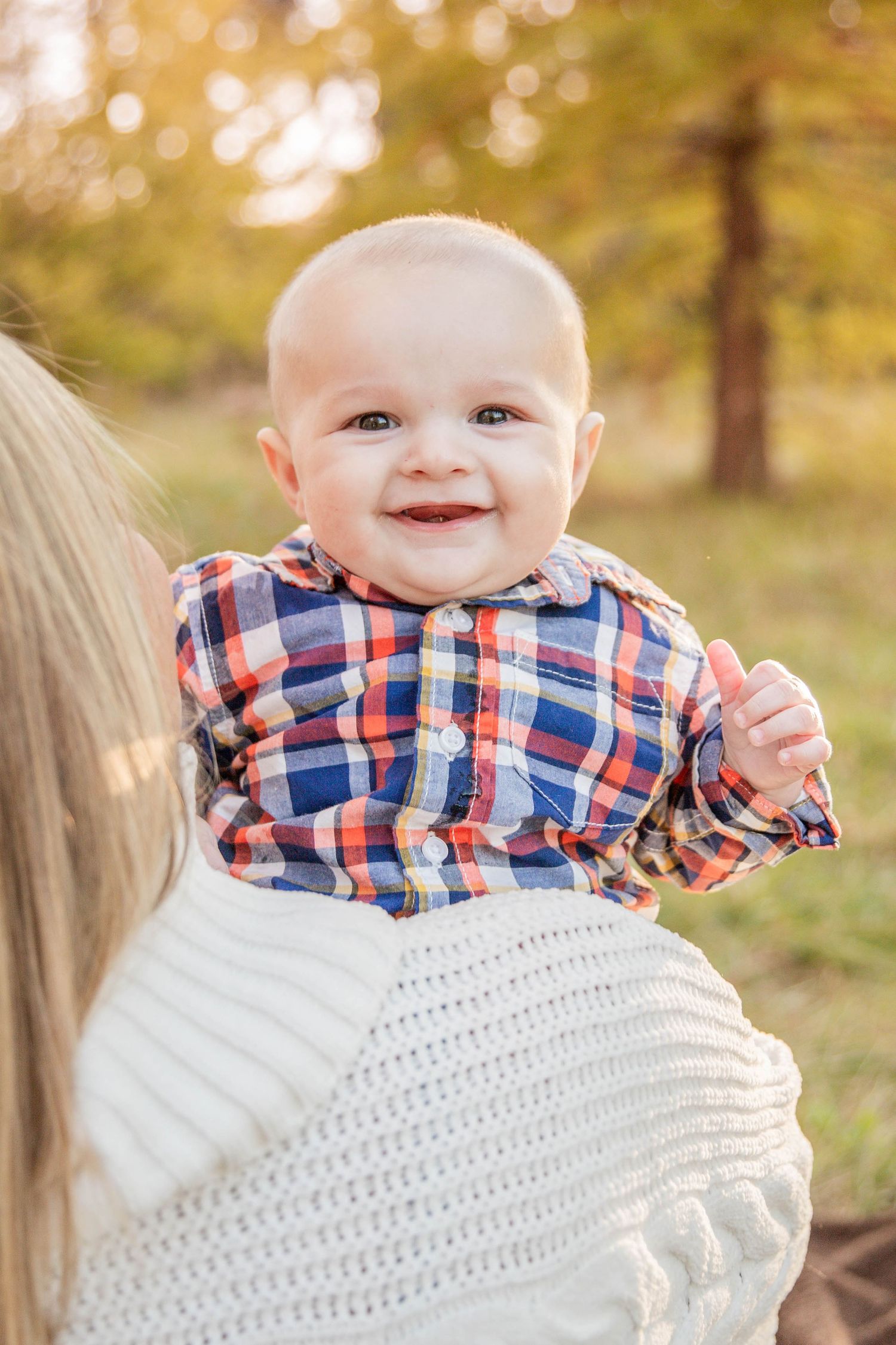 Baby in plaid shirt smiles while being held outdoors in autumn setting.
