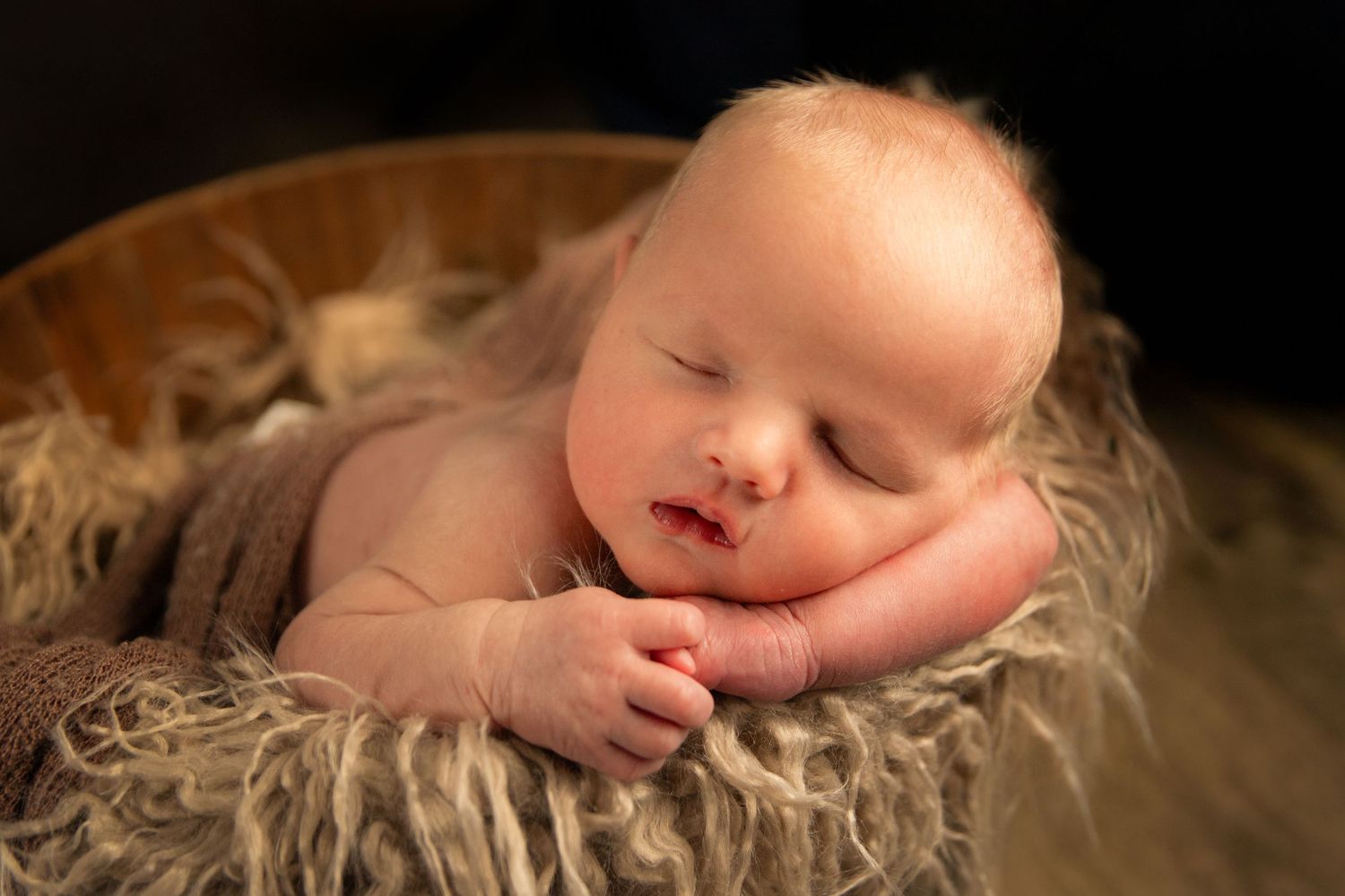 Peaceful newborn baby sleeping in a wooden bowl lined with soft fur, hands tucked under chin.