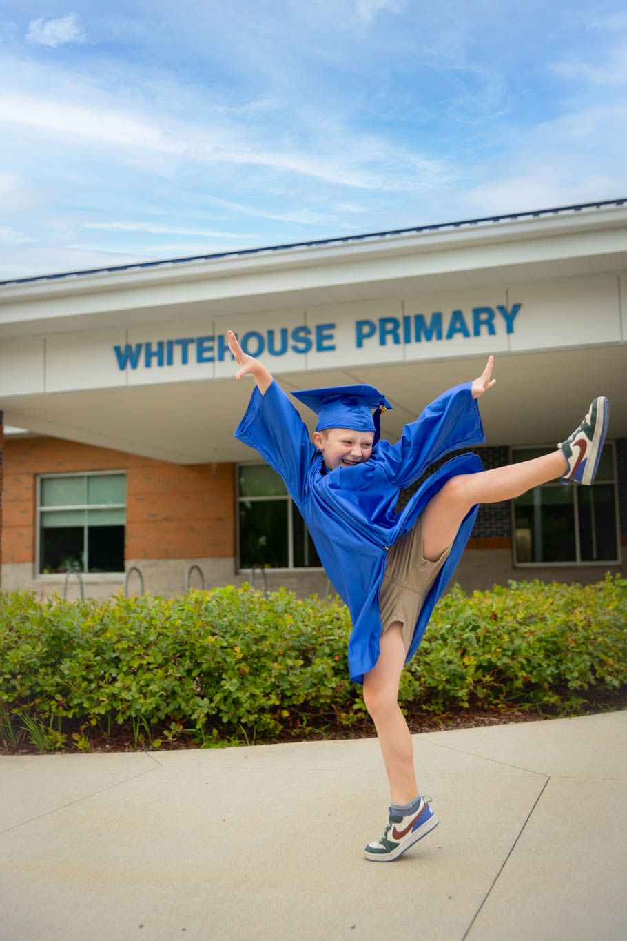 Graduate in blue cap and gown kicks leg high in front of Whitehouse Primary school.