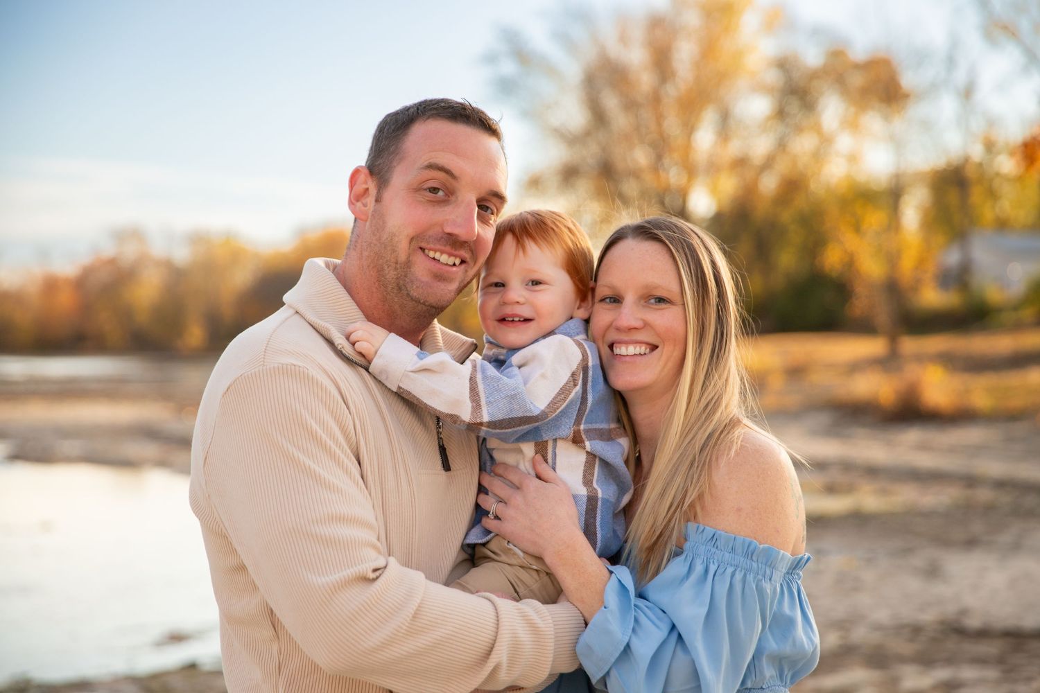 A family wearing coordinated light blue and beige outfits embraces during a golden sunset photoshoot outdoors.