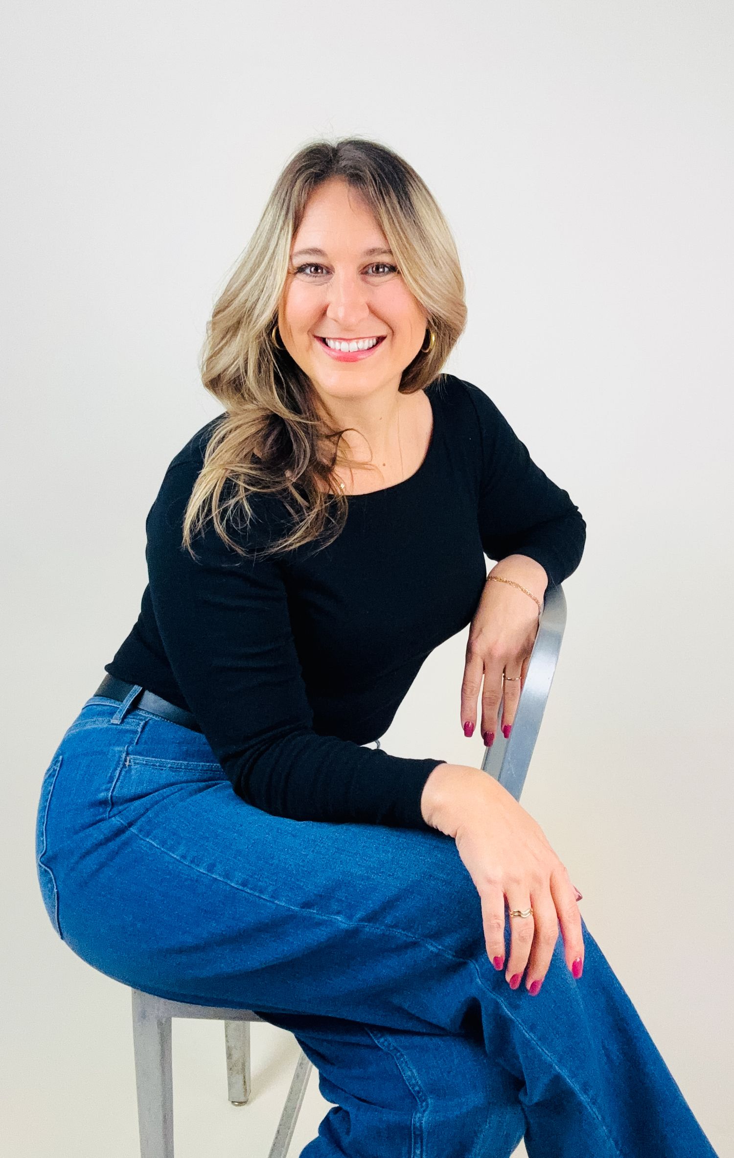 Woman in black sweater and blue jeans poses casually on white chair in studio setting.