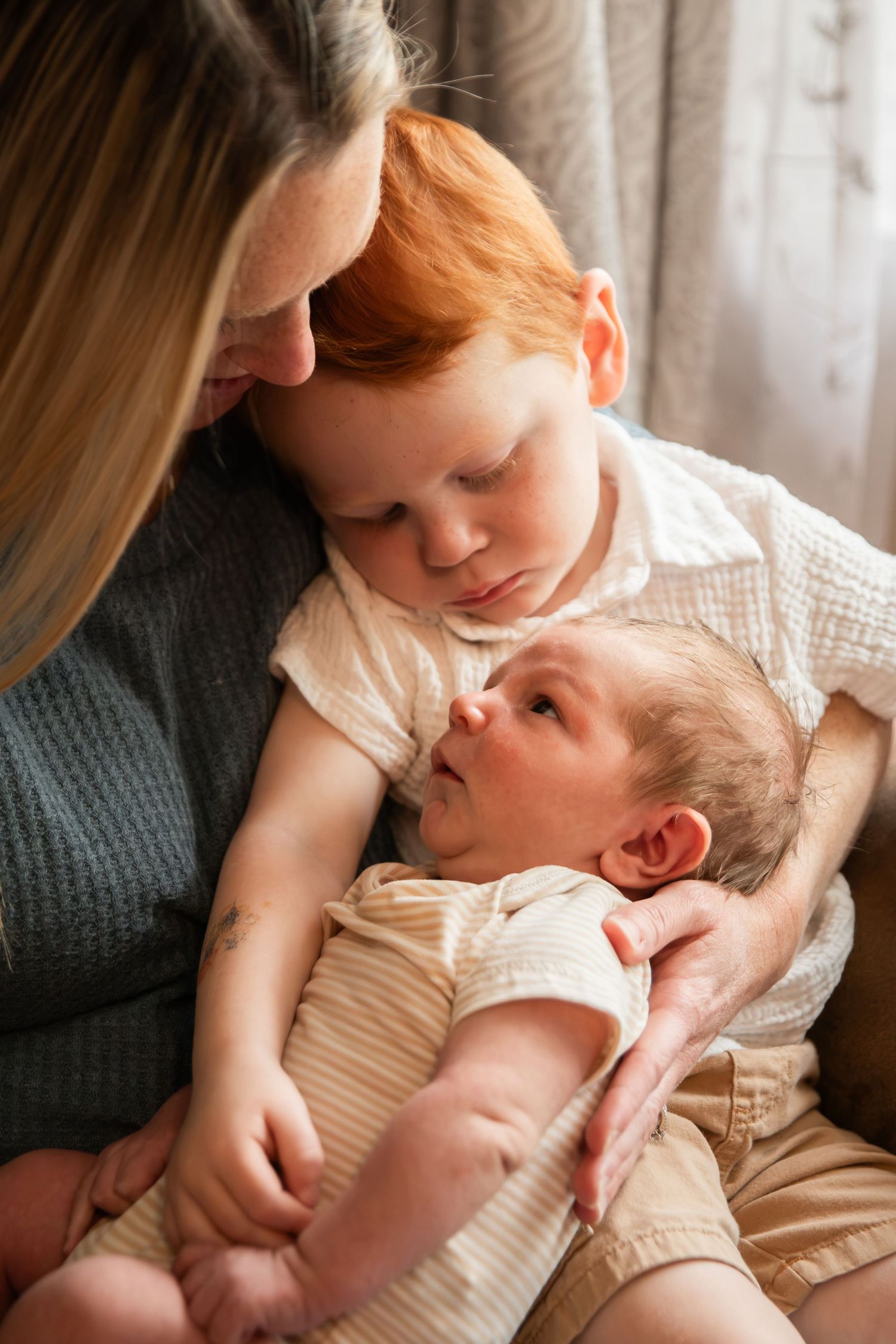 A mother holds her newborn baby while her young red-haired toddler gently leans in to look at the infant.