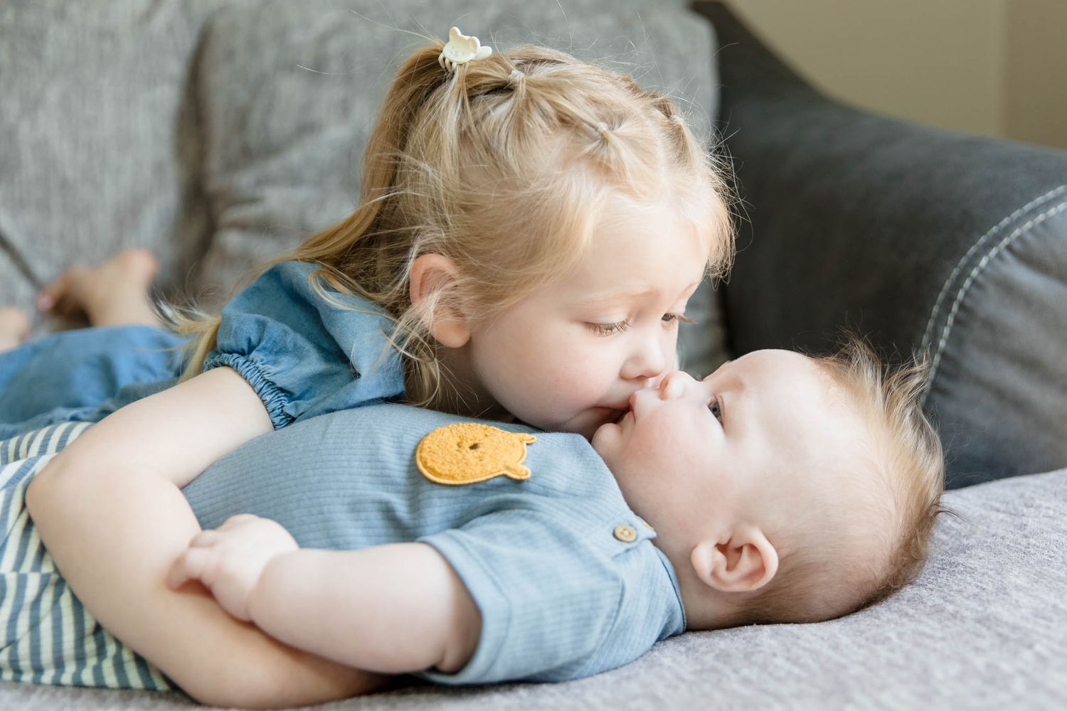 Two children share tender moment while resting on gray couch.