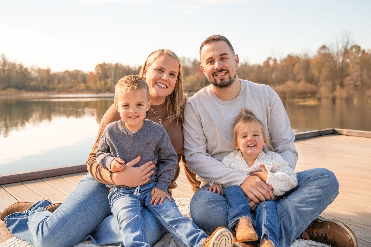 A family sits together on a wooden dock by a lake during golden hour, wearing casual blue clothing.