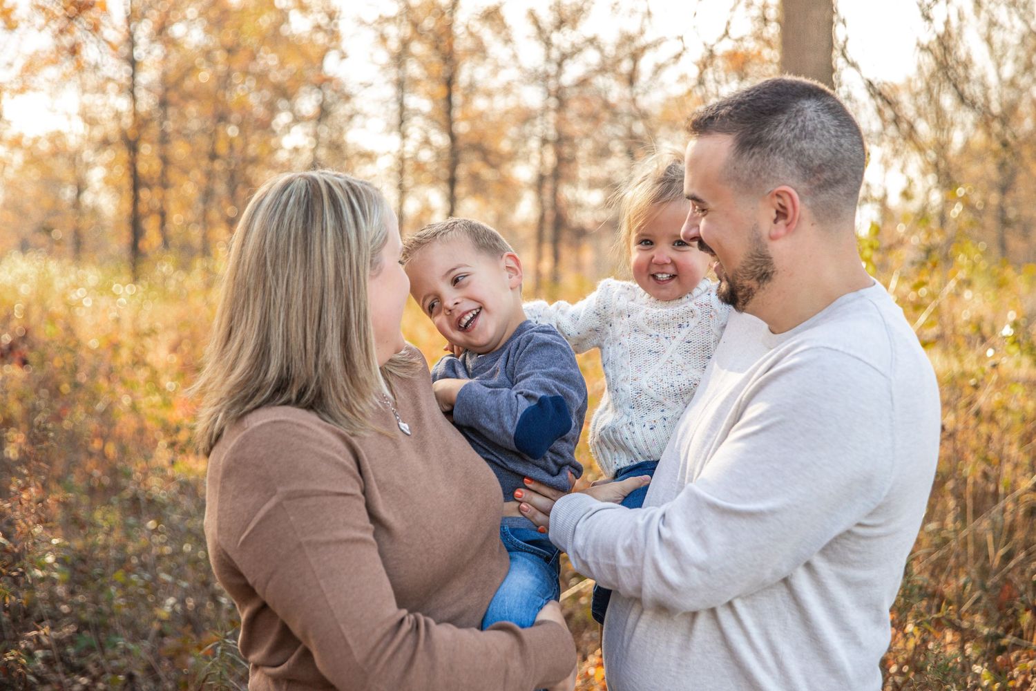 A family shares joyful laughter together during an outdoor autumn photoshoot with golden foliage in the background.