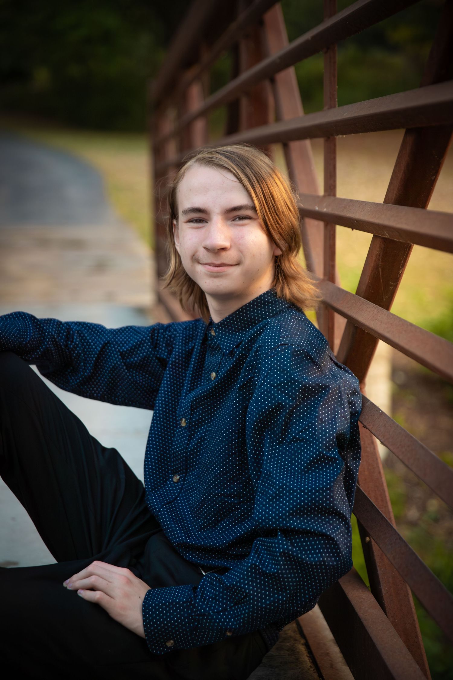 Outdoor portrait of someone in a navy polka dot button-up shirt sitting casually against a wooden bridge railing.