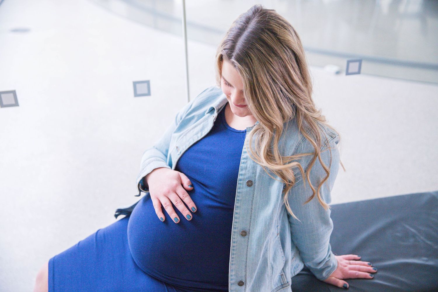Pregnant woman in denim jacket rests hand on belly while sitting in medical office.