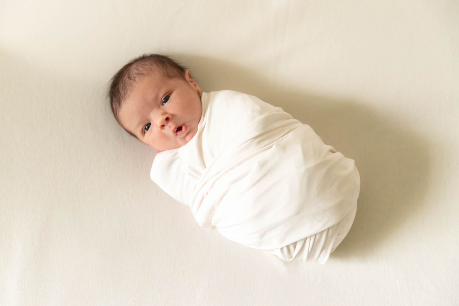 Newborn baby swaddled in white wrap on cream background.