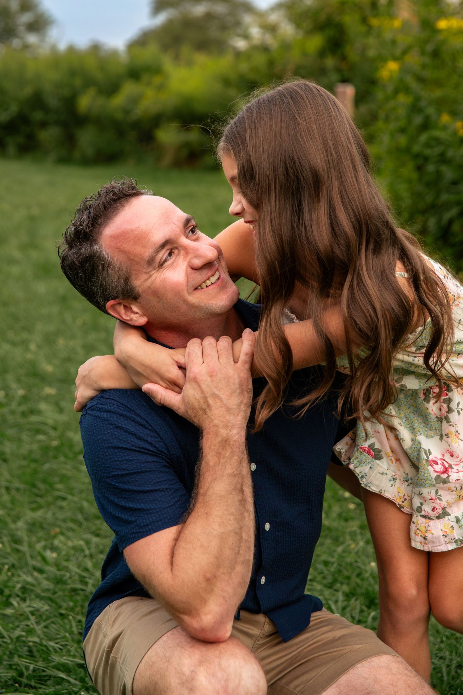 A young girl kisses her smiling father on the cheek while embracing outdoors on green grass.