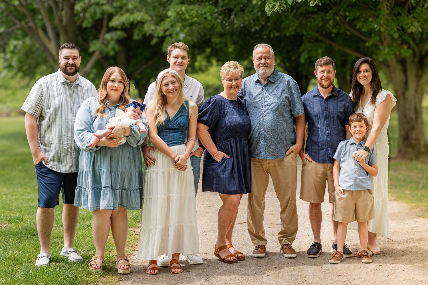 A multigenerational family of eight poses together outdoors on a sunny day, smiling warmly on a tree-lined path.