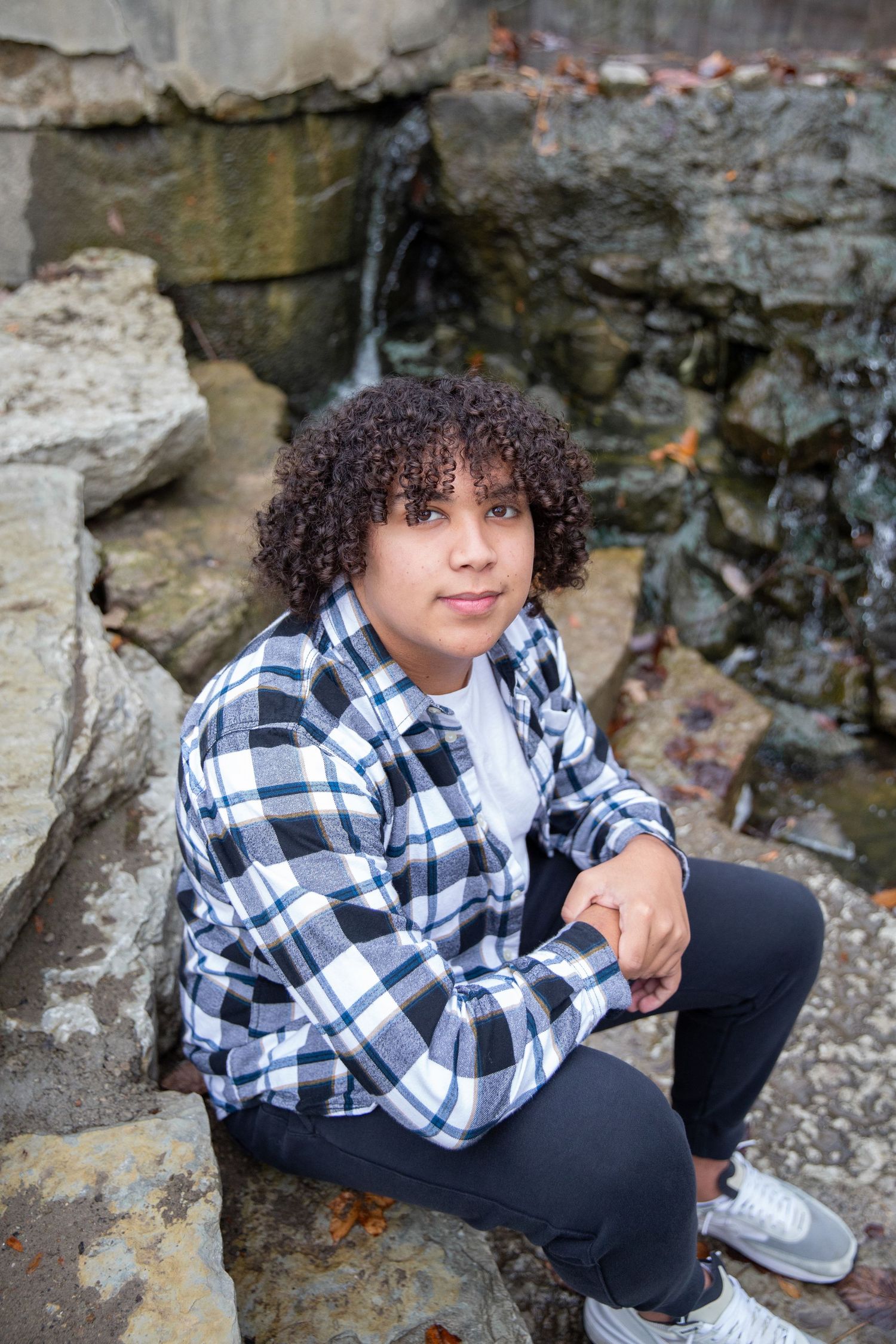 Person in plaid flannel shirt sitting on rocky outcrop near waterfall.