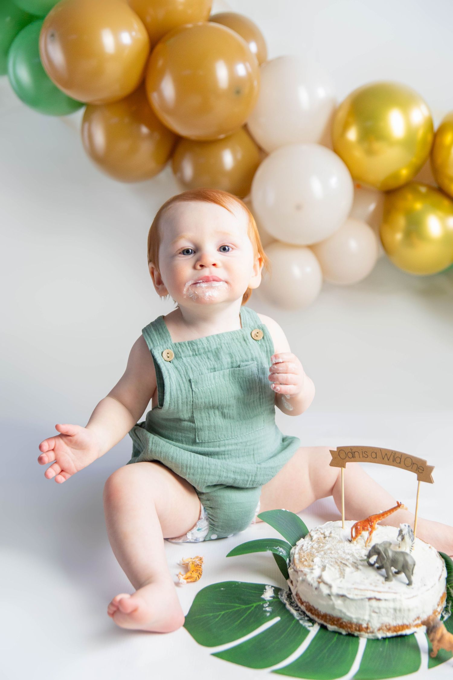 Baby in mint green romper sits by balloon arch and birthday cake on tropical leaf.