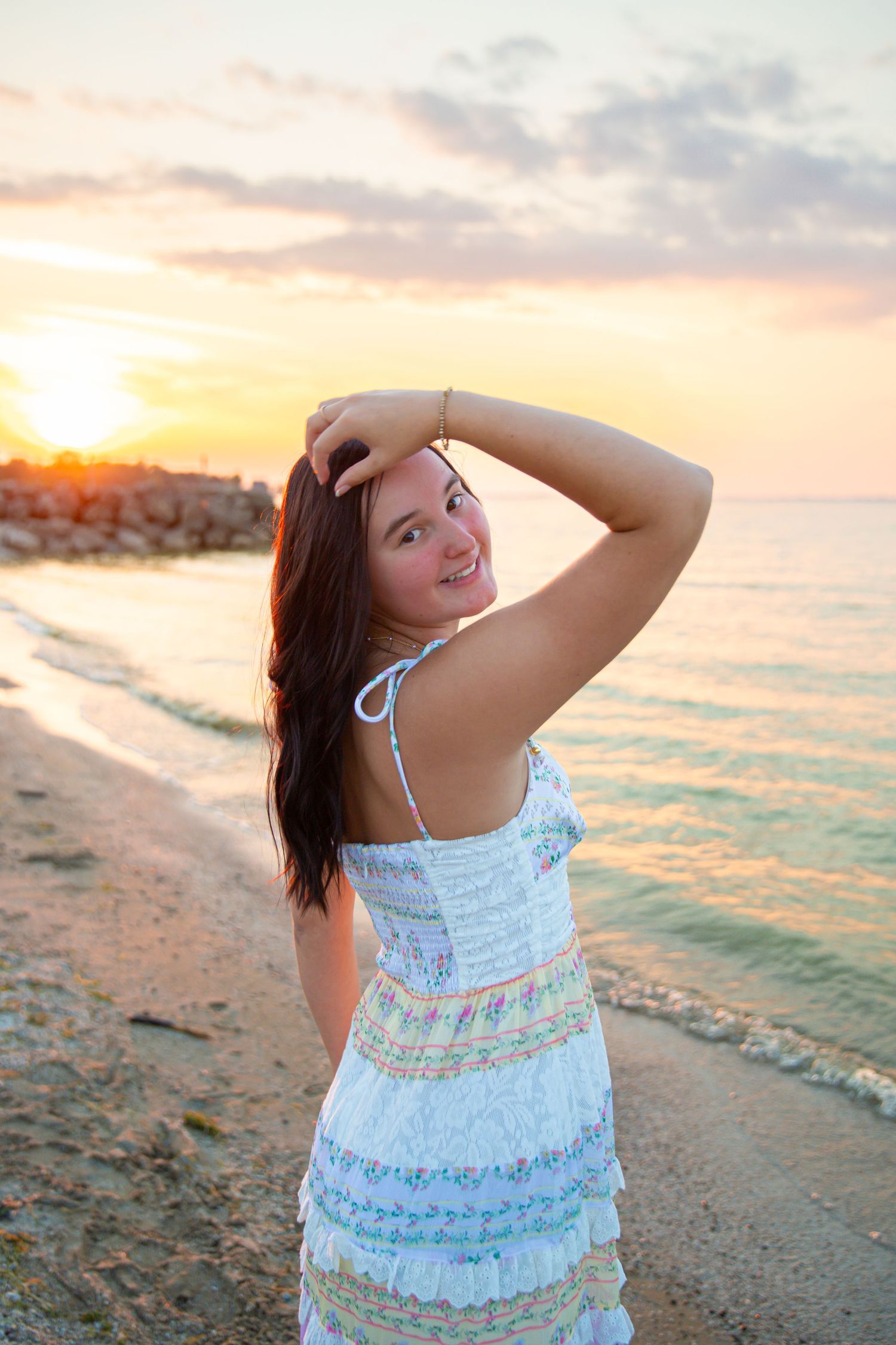 Young woman in white sundress poses on sandy beach at sunset, smiling over shoulder with hand in dark hair.