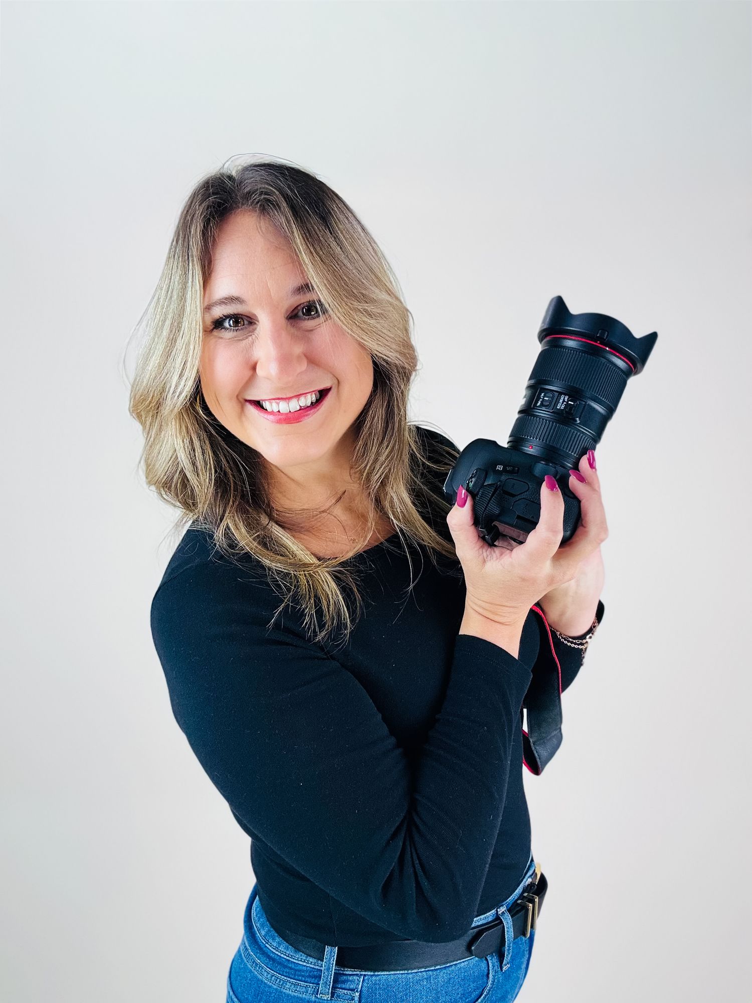 Professional photographer wearing black top and jeans holds camera while posing against white background in studio.